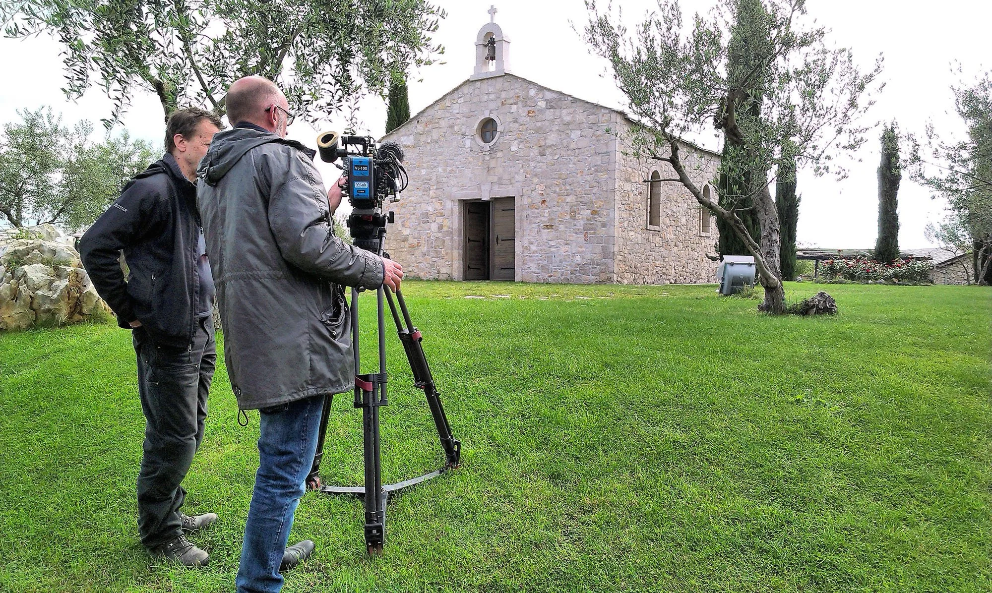 Two men filming a small stone church with a cross on top, amid trees and green grass.