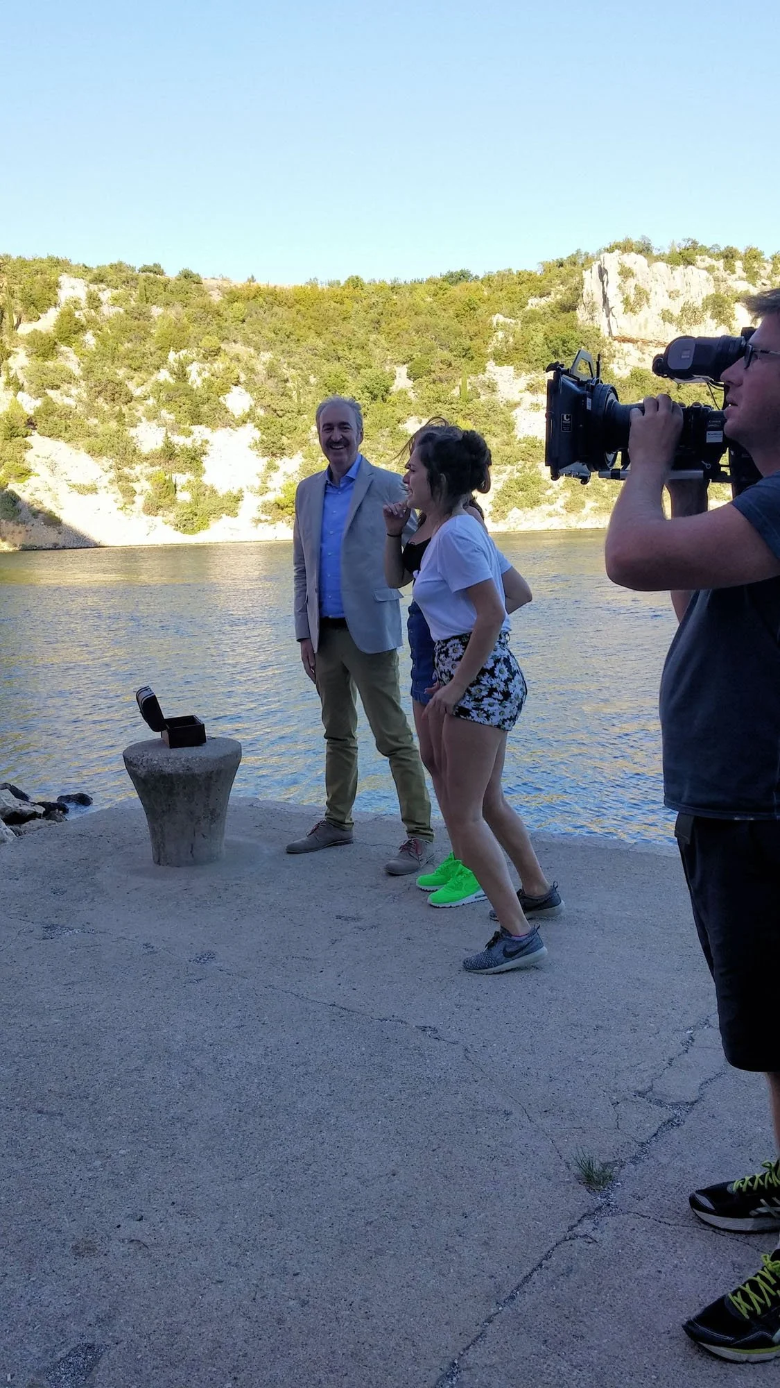 A group of people filming a scene outdoors near a body of water with a rocky hillside and trees in the background. One person is holding a professional video camera, and another two people are standing and smiling.