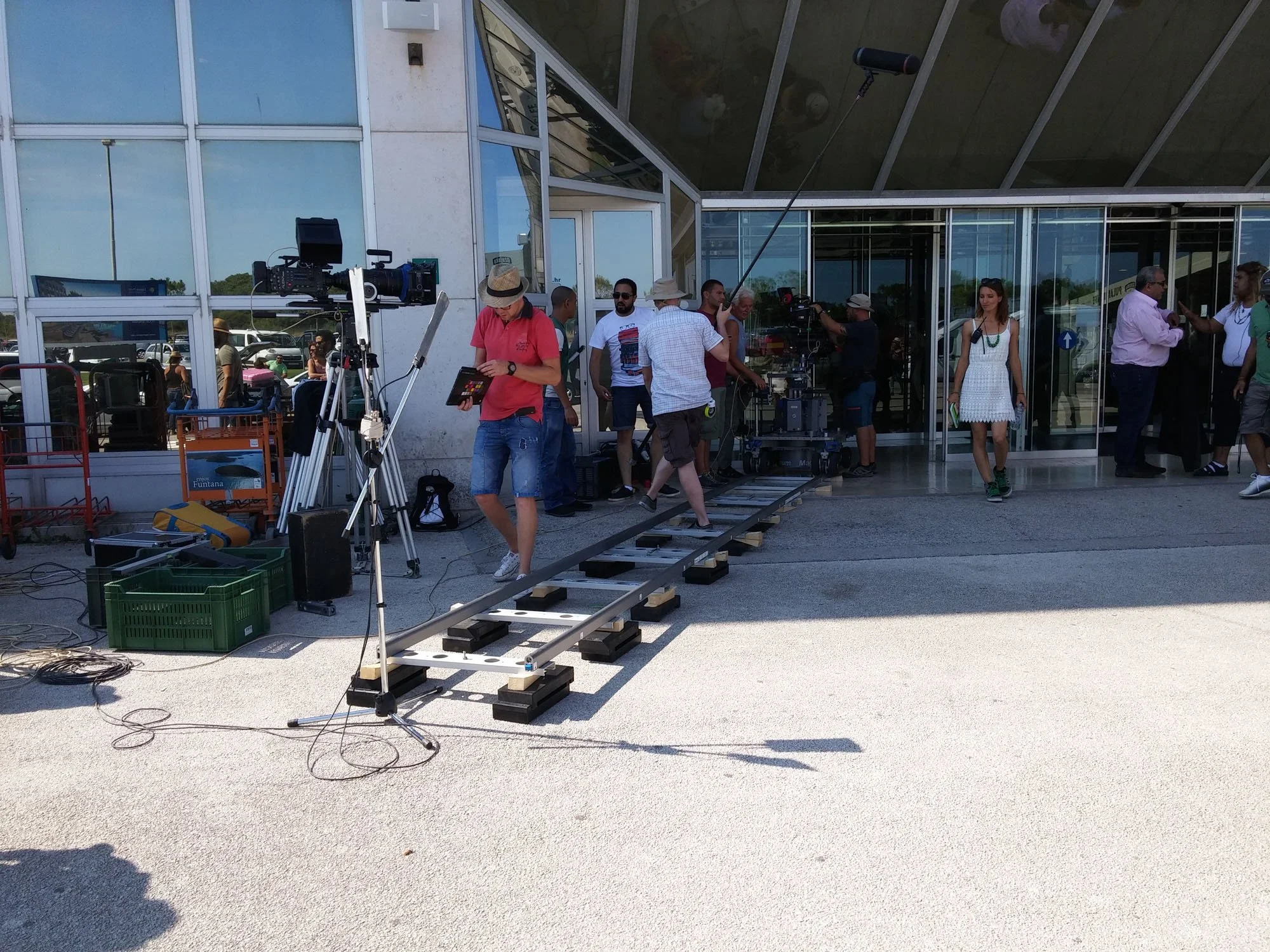 Film crew setting up camera equipment outside a modern building with large glass windows. Crew members are preparing a camera crane on rolling wheels, with additional electronic gear and carts nearby.
