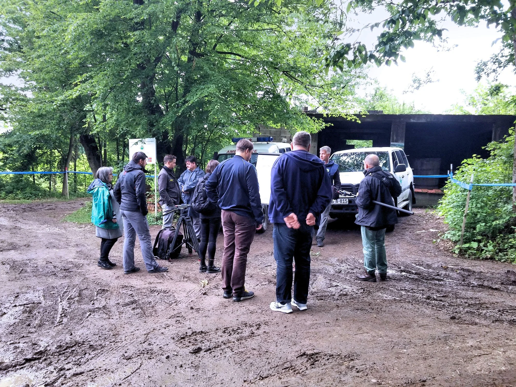 Group of people, including police officers and civilians, gathered outdoors near police vehicles under green trees, with an emergency sign and a dark tunnel or underpass in the background.