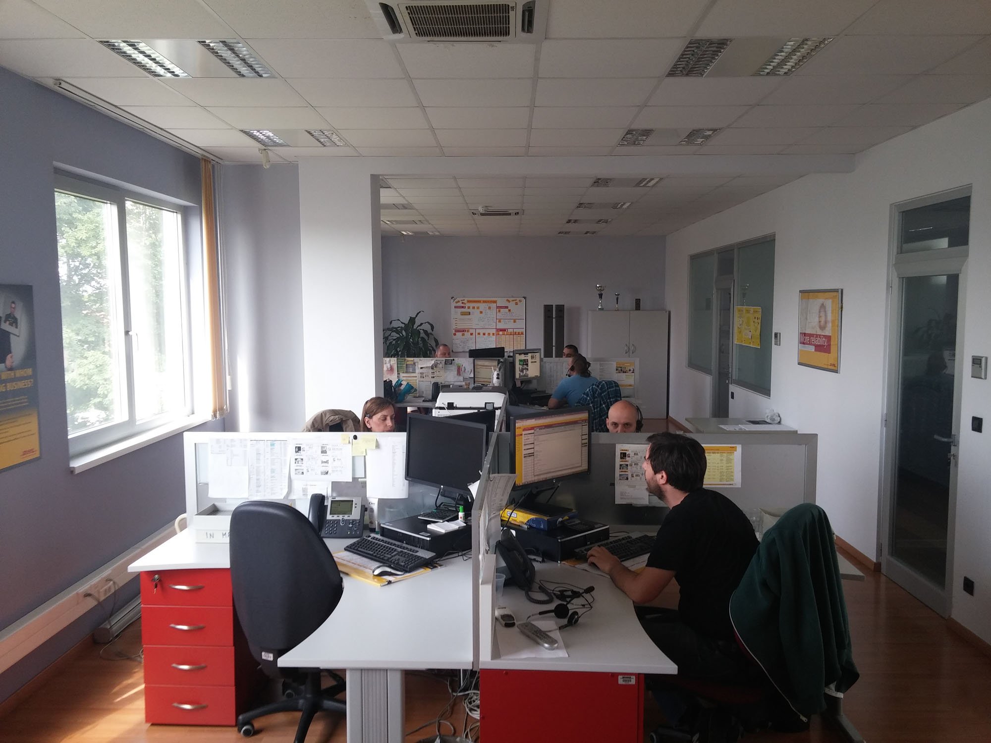 Open-plan office with employees working at desks with computers, some engaged in conversation, and large windows letting in natural light.