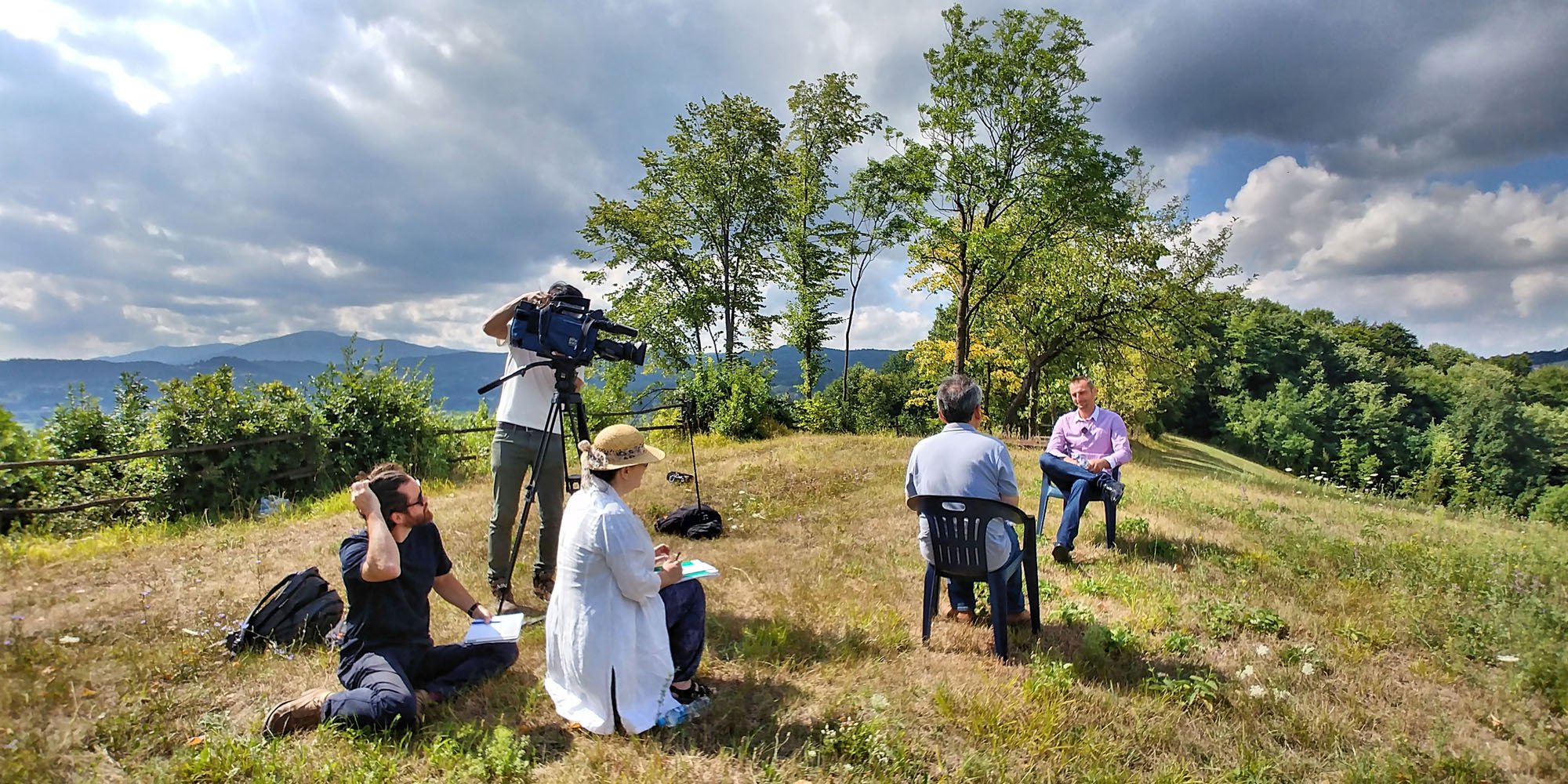 A group of five people outdoors in a grassy field with trees and mountains in the background, some seated and one standing, while a camera operator films a man in a purple shirt sitting on a chair, possibly during an interview or documentary filming.
