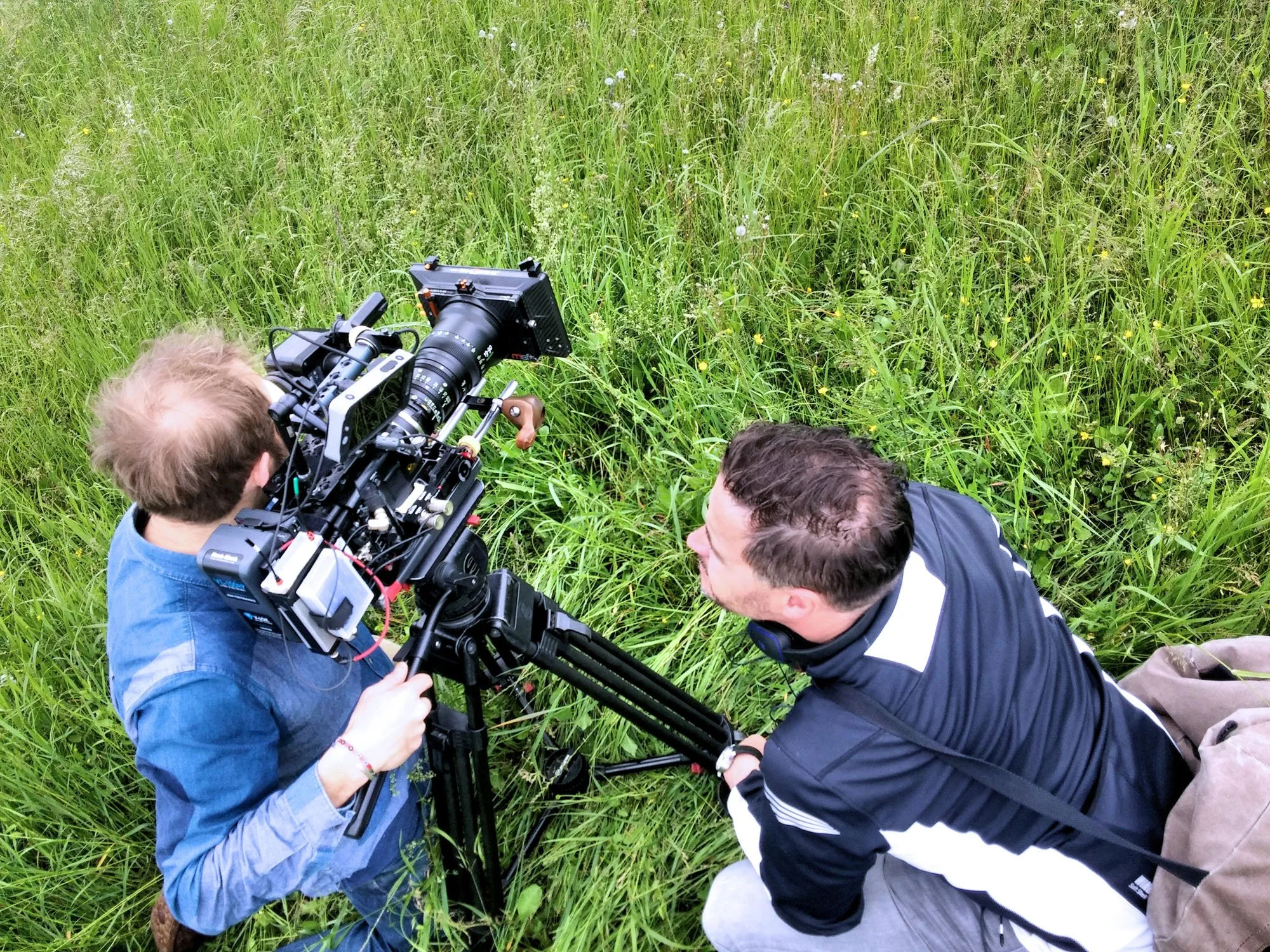 Two men operating a professional camera on a tripod in a grassy field with tall green grass and small yellow flowers.