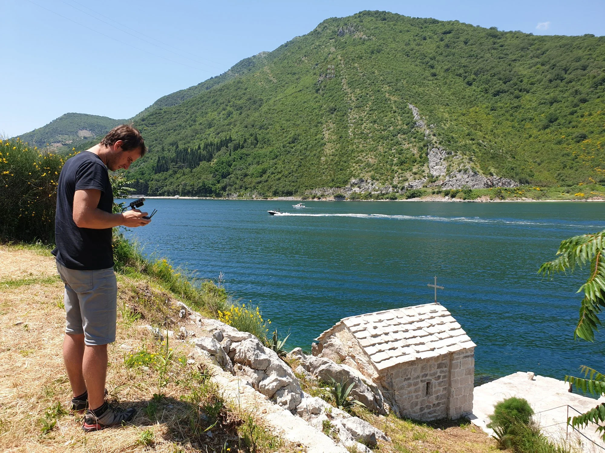 A young man wearing a black t-shirt, gray shorts, and black hiking shoes stands on a rocky path by a lake, filming with drone, aerial. Behind him, lush green hills and mountains rise up, and a boat moves across the water. The sky is clear and sunny.