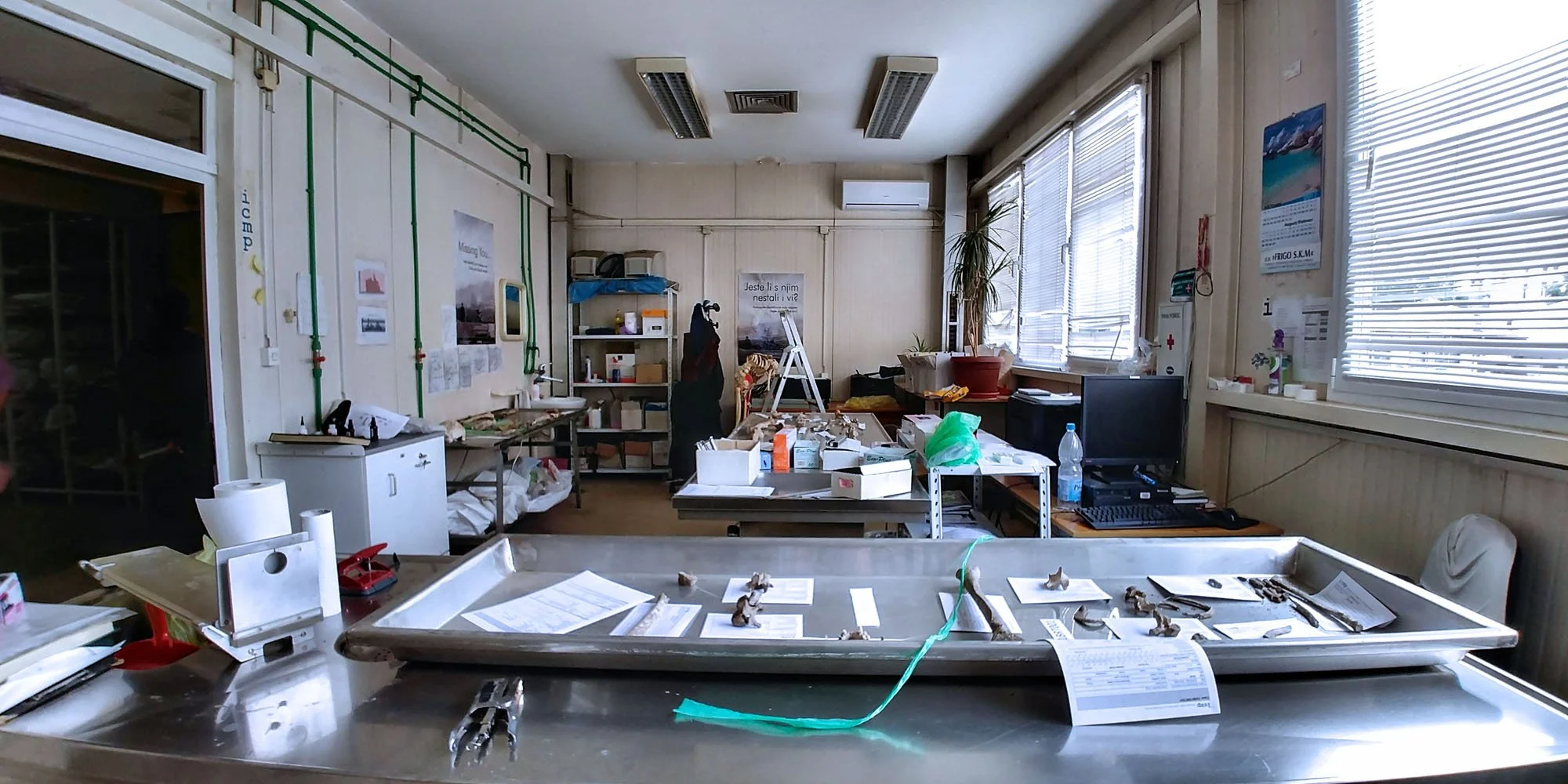 A cluttered office or medical workspace with tables, shelves, and equipment. Items include papers, boxes, a water bottle, and medical or lab instruments.