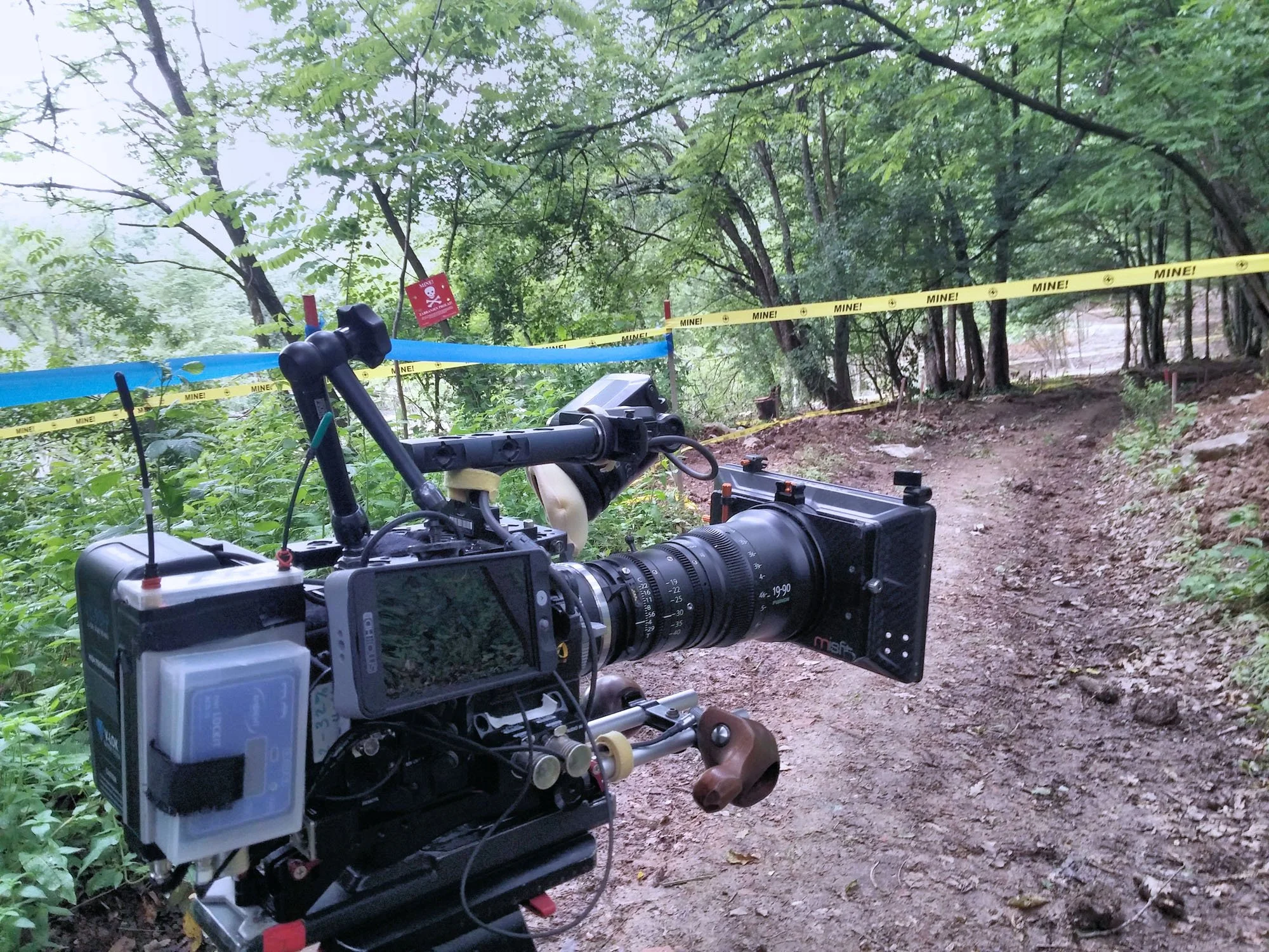 A professional camera mounted on a tripod capturing footage in a wooded outdoor area marked off with yellow 'MINE!' tape and red warning signs, suggesting a scene of a crime or mine-related activity.
