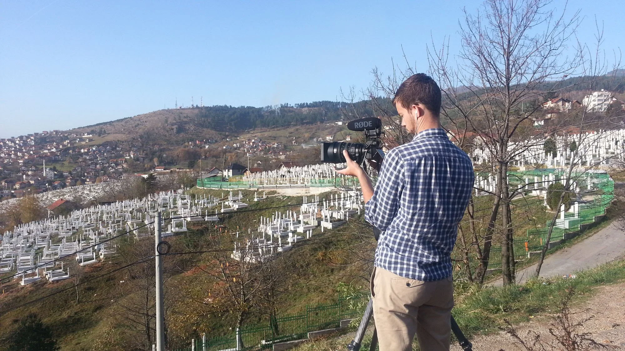 A man filming outdoors with a professional camera in a hilly area with numerous white crosses on the ground, a residential neighborhood on the hillside, and leafless trees in the foreground.