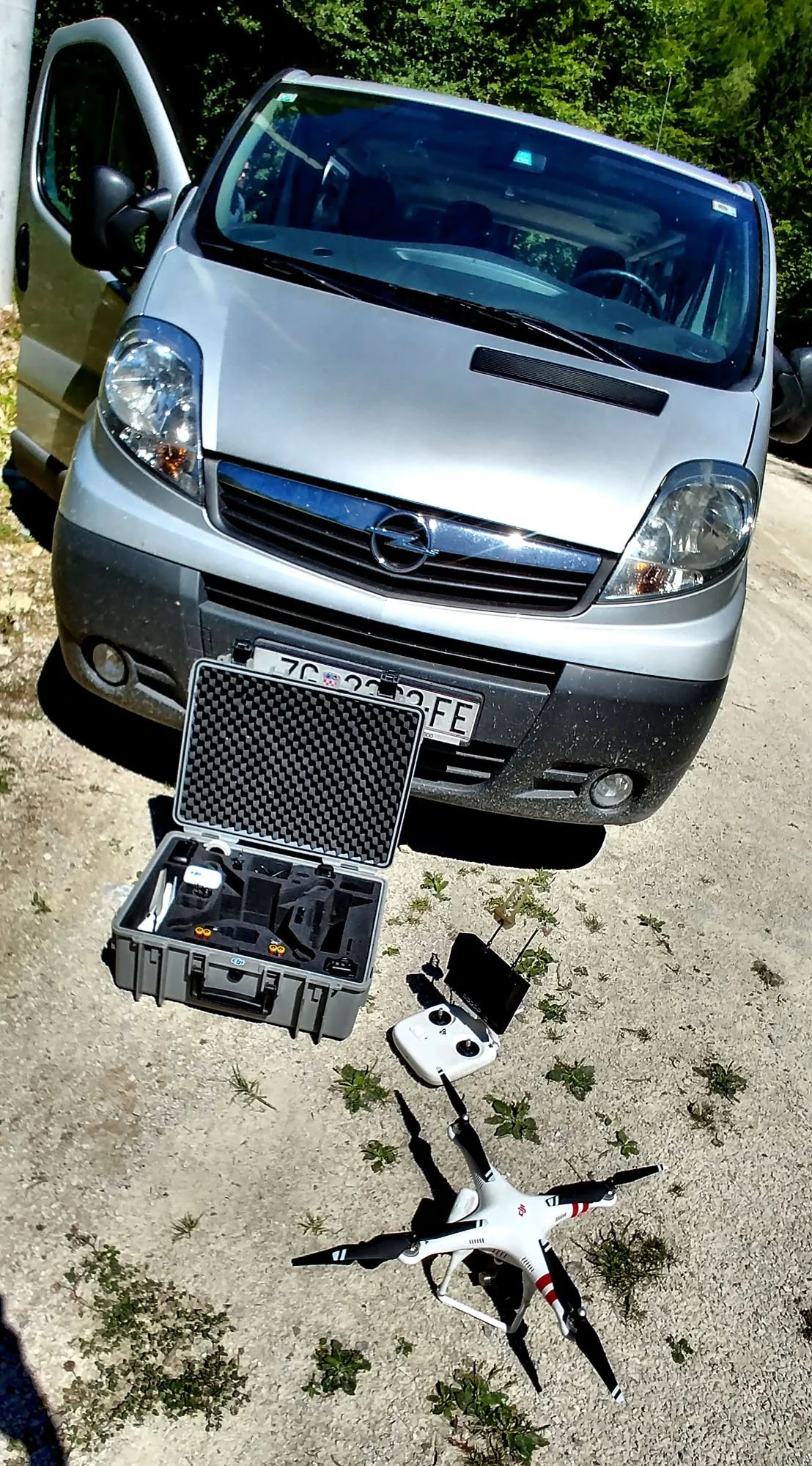 A silver Opel van with open side door parked on a dirt surface, with a drone, remote controller, and a protective case laid out in front of it, all outdoors with greenery in the background.
