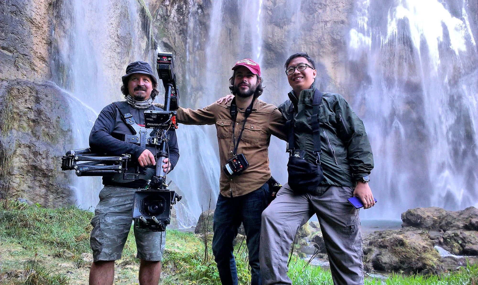 Three men standing together outdoors in front of a waterfall, smiling and posing for a photo. The man on the left is holding camera equipment, the man in the middle has a camera around his neck, and the man on the right is holding a phone.