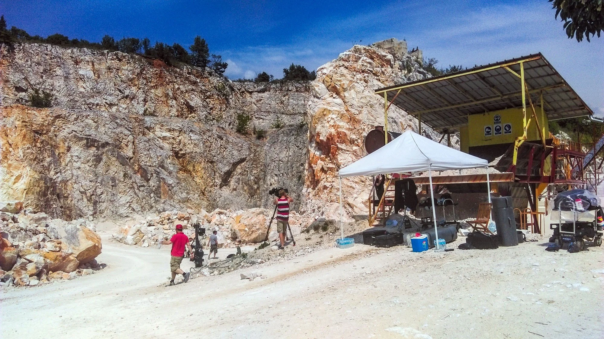 Filming crew working at a quarry site with rocky cliffs and a large yellow filming platform under a tent, with equipment and personnel scattered around.