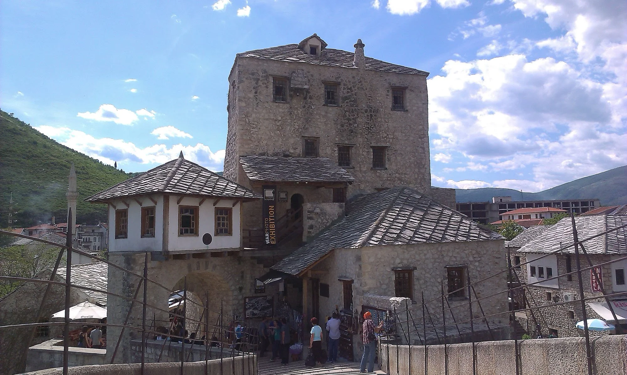 A historic stone tower building with small windows, surrounded by buildings and hills, with a crowd of people walking underneath and a sign that reads 'War Photo Exhibition'.