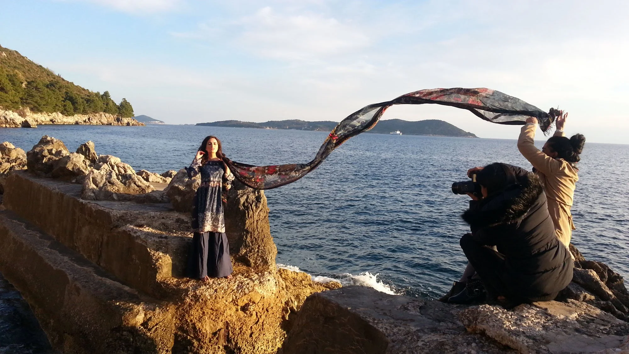 A woman in a long dress is being photographed by a photographer on a rocky shoreline near the water, with a scenic view of islands and a boat in the background. Another person is holding a large, flowing scarf to create a dynamic effect.