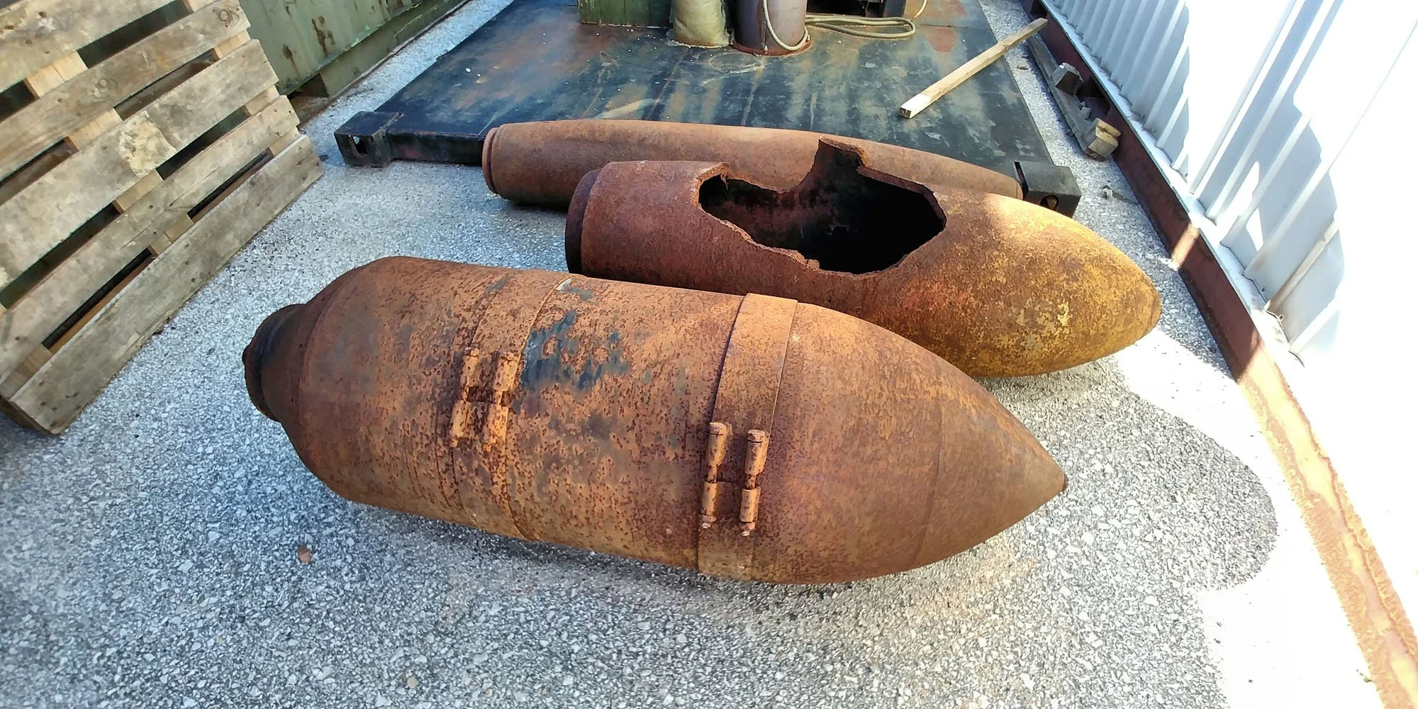 Three large, rusted bombs lying on a gravel surface next to a wooden pallet and a metal fence.