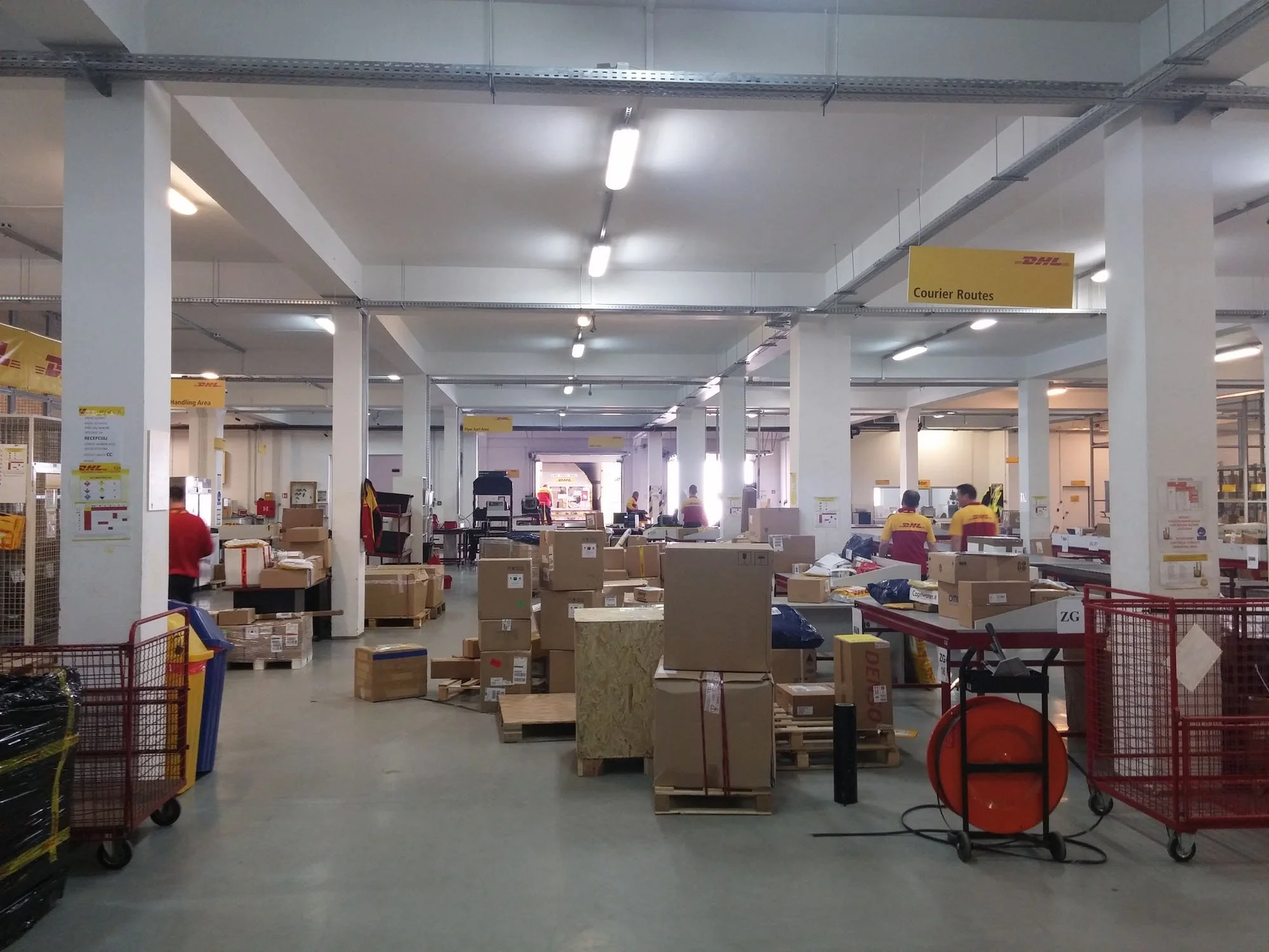 Inside a DHL courier sorting facility with workers managing parcels, surrounded by cardboard boxes and carts, under white ceiling lights.