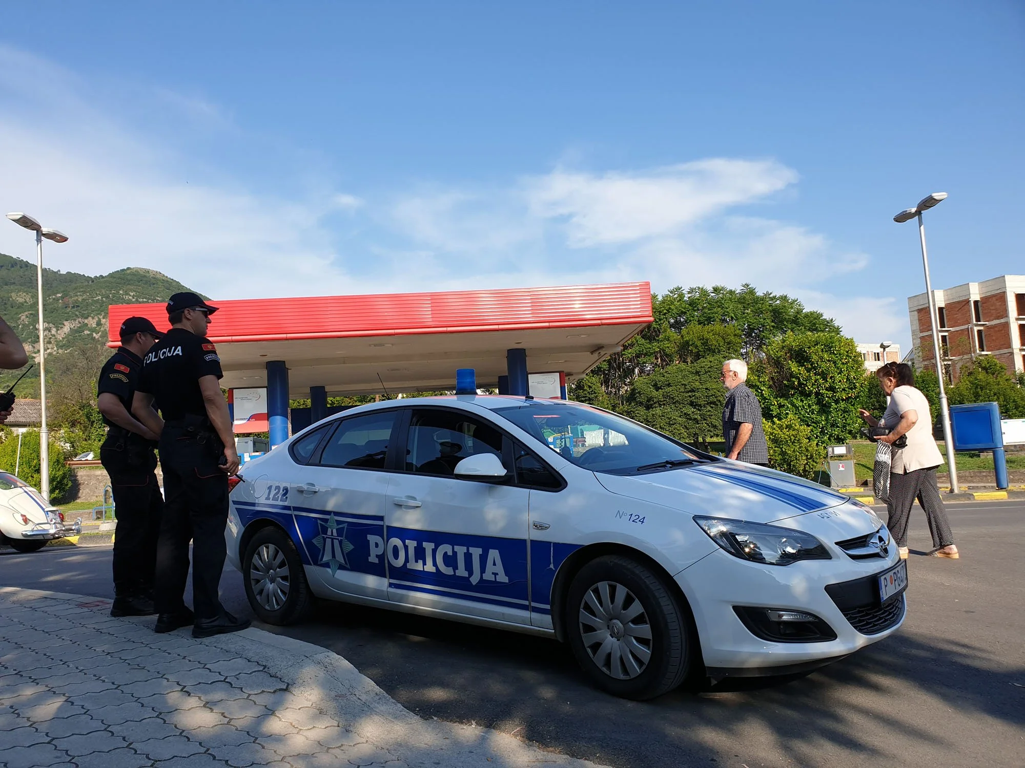 Police officers and civilians gathered around a police car marked 'Policija' near a gas station, with a mountain and a building in the background, during daytime.