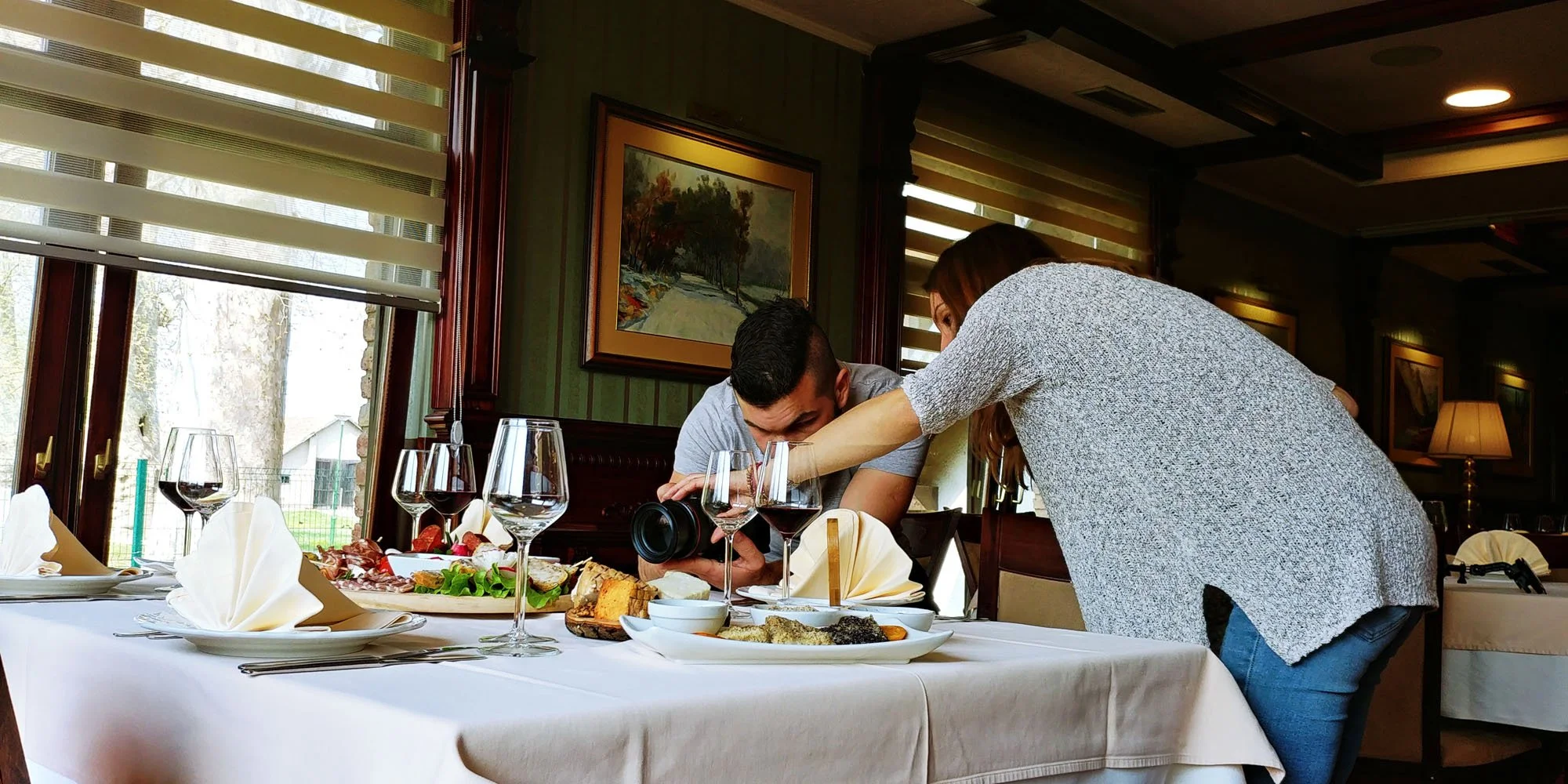 A woman leaning over a man, taking a photo of a food spread at a restaurant table with wine glasses, bread, and salads.