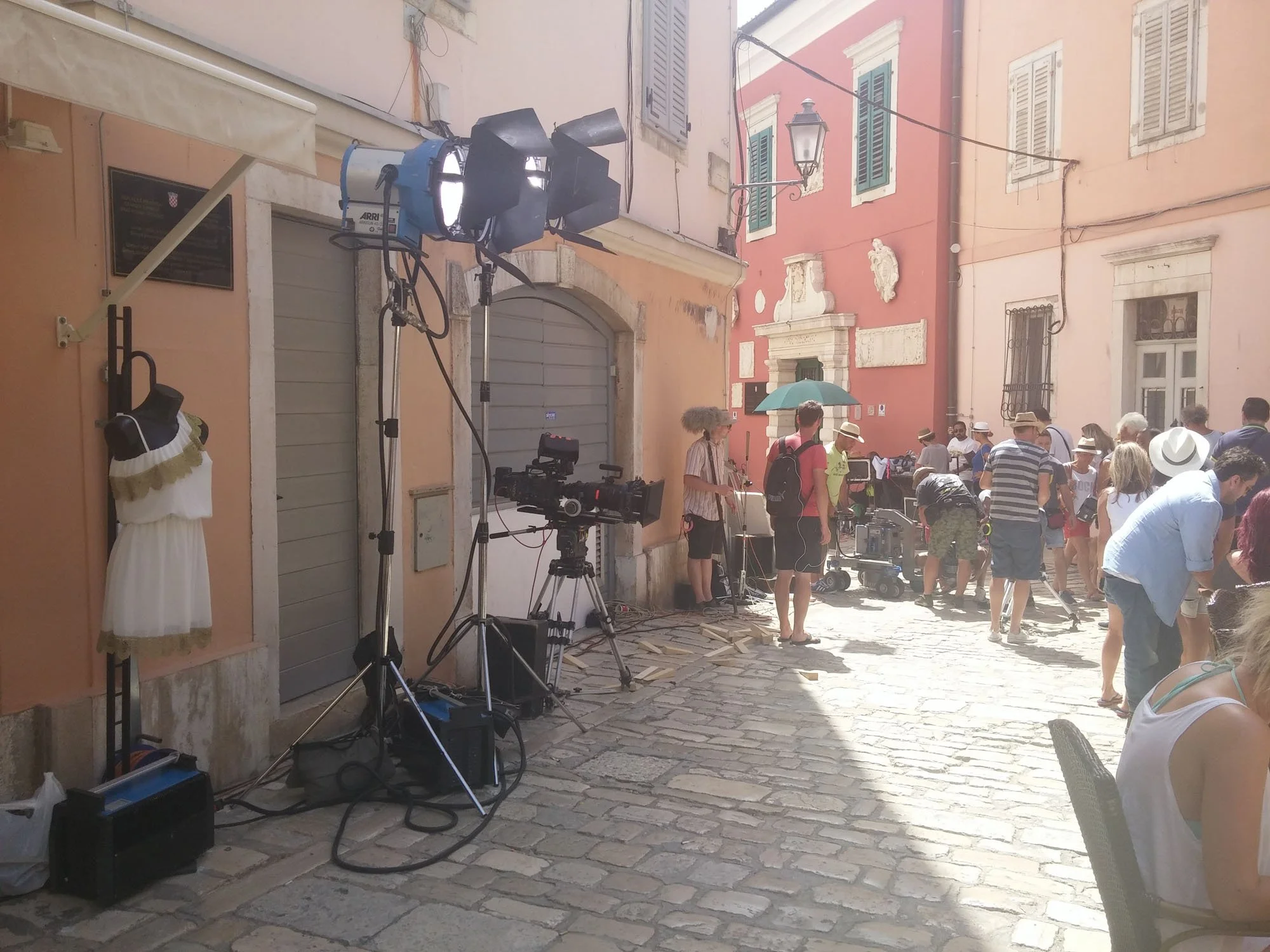 Outdoor scene at a film or photo shoot with lighting equipment on a rustic cobblestone street, surrounded by pastel-colored buildings with shuttered windows, a crowd of people including crew members and onlookers, one person holding an umbrella, and 