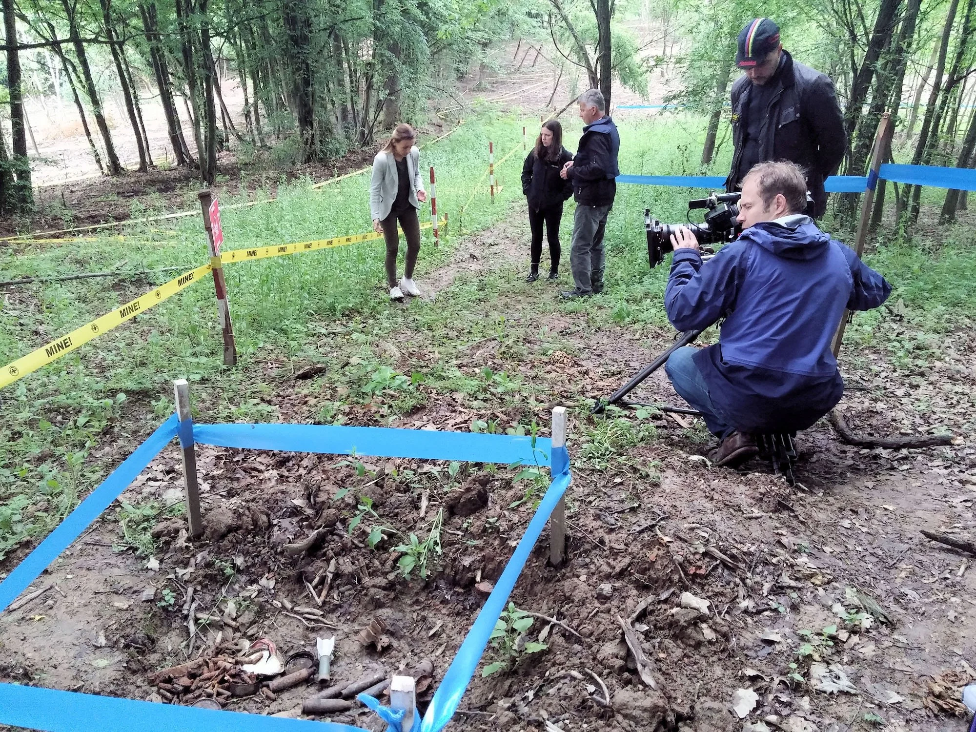 Group of people inspecting an outdoor archaeological site or excavation in a wooded area, with blue and yellow markings and caution tape around the area, and a person filming the scene.