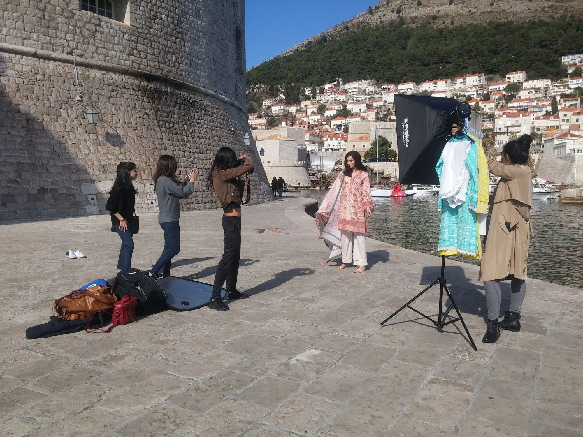 A photoshoot setup beside a harbor with a city hillside in the background. Two women are posing for photos, one in a pink dress, surrounded by three women taking pictures and adjusting a large softbox lighting equipment. Luggage and bags are placed o
