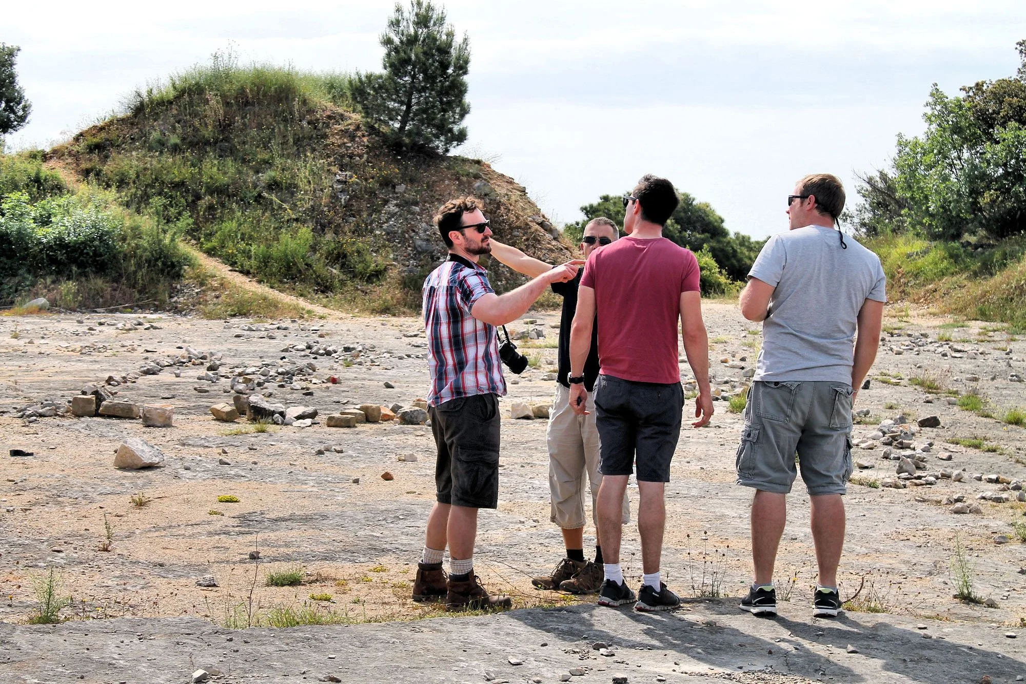 Group of five men standing and talking on a rocky outdoor area with a small hillside, trees, and shrubs in the background.