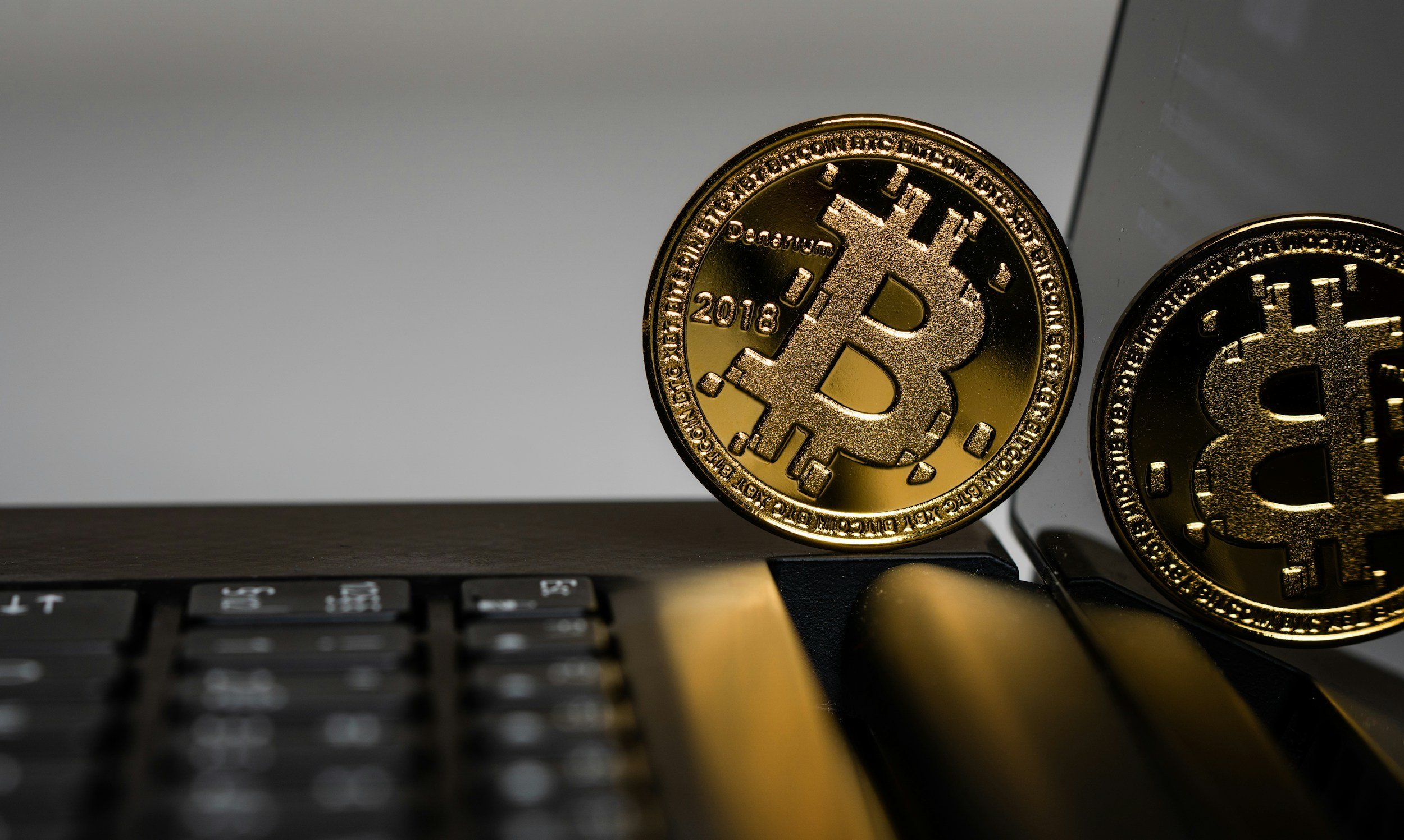 Close-up of two gold-colored Bitcoin coins, one leaning against a reflective surface and the other on a black keyboard, with a blurred background.
