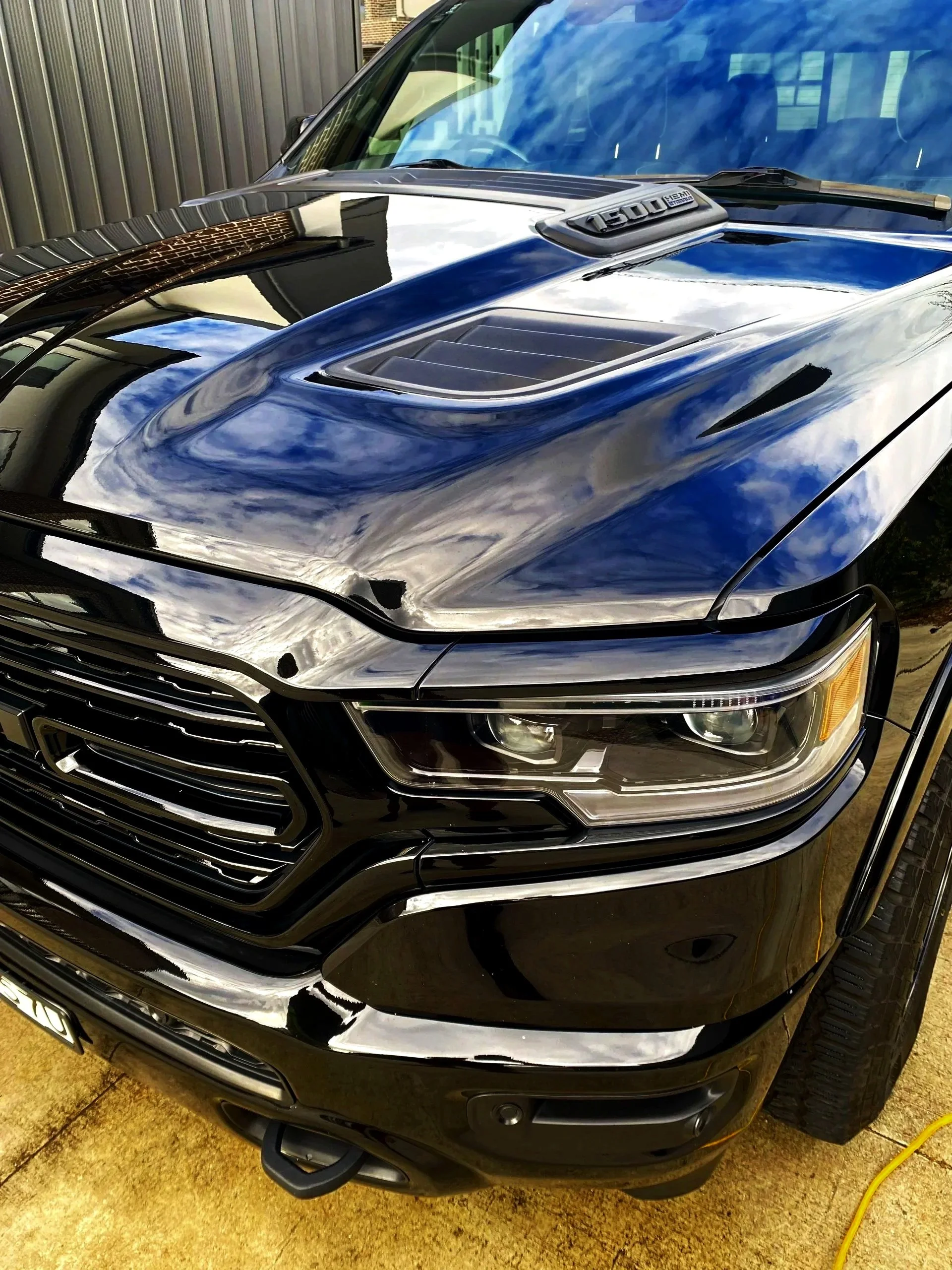A black luxury SUV with a shiny exterior reflecting the sky and clouds. The front grille and headlight are visible, along with a hood vent and part of the windshield. The vehicle is parked on a concrete surface.