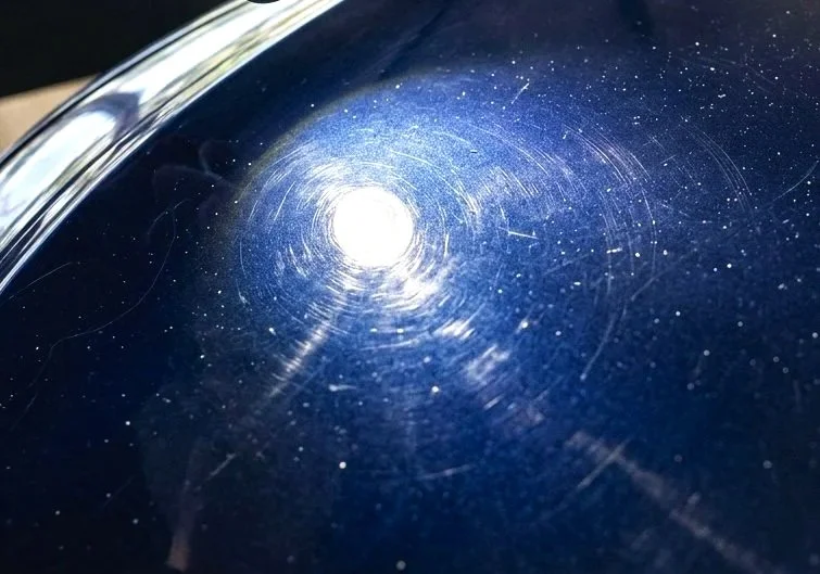 Close-up of a dark blue car surface with visible swirl marks and a bright reflection of light.