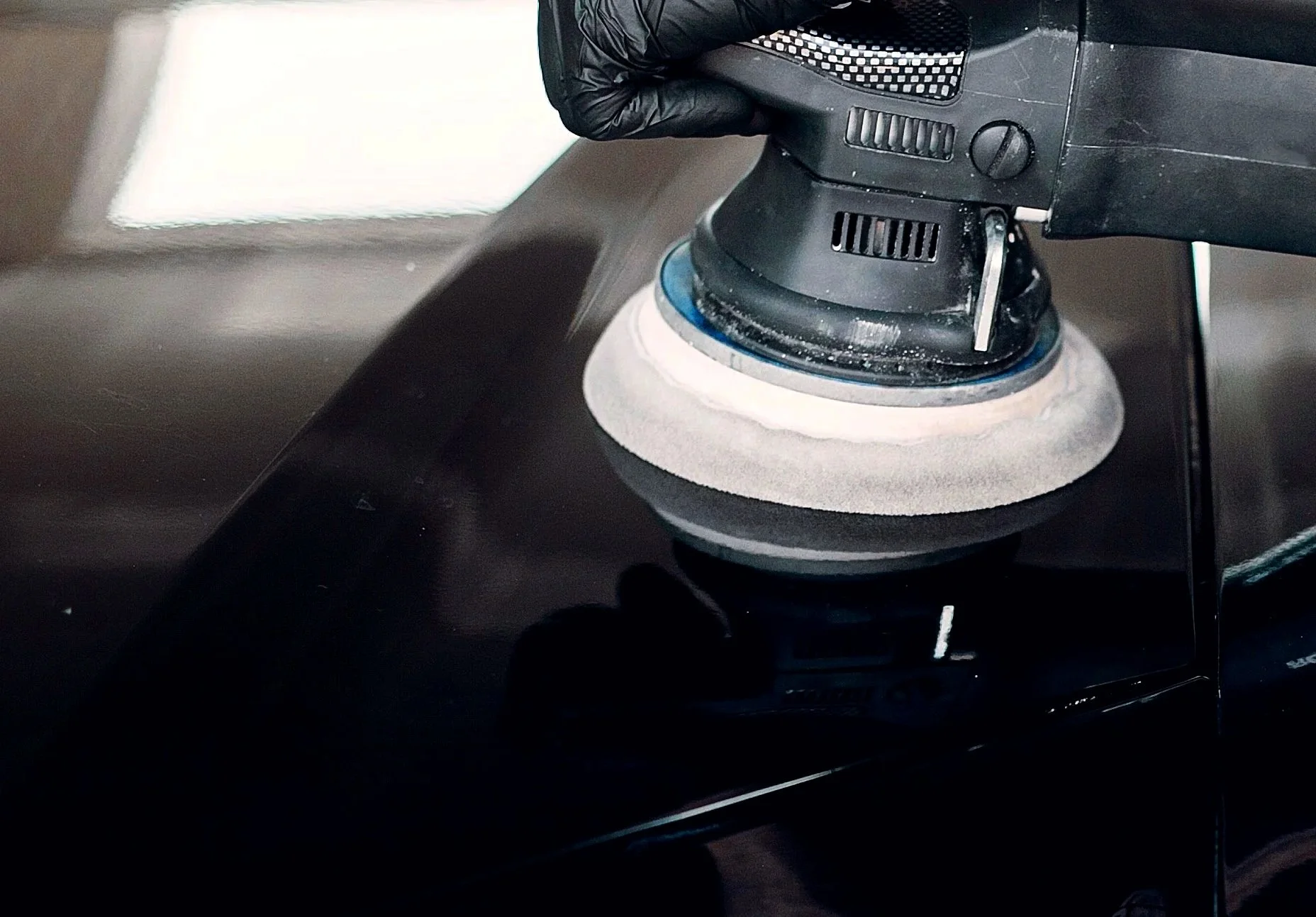 Close-up of a black electric polisher with a white foam pad, being used on a black vehicle surface, operated by Ty.