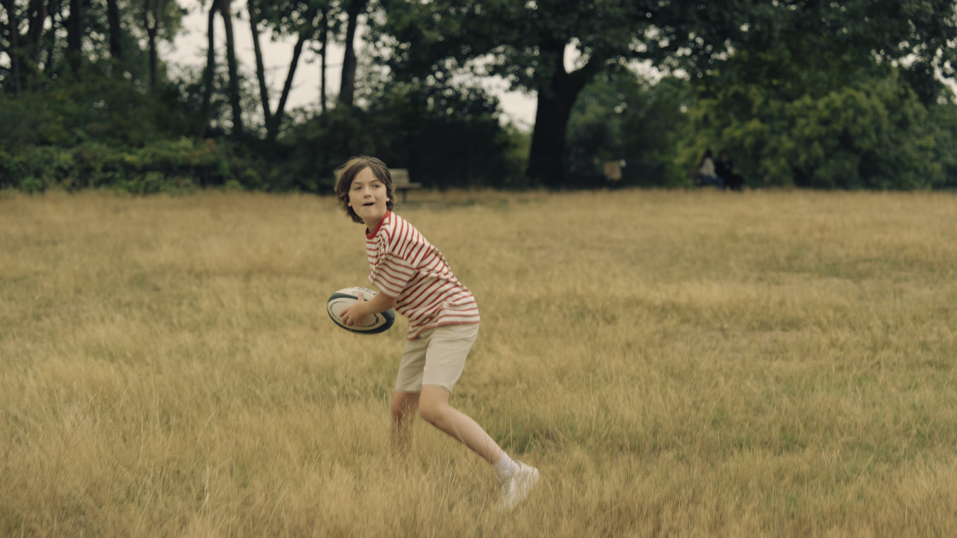 A young boy playing rugby on a grassy field, holding a rugby ball and looking to the side.