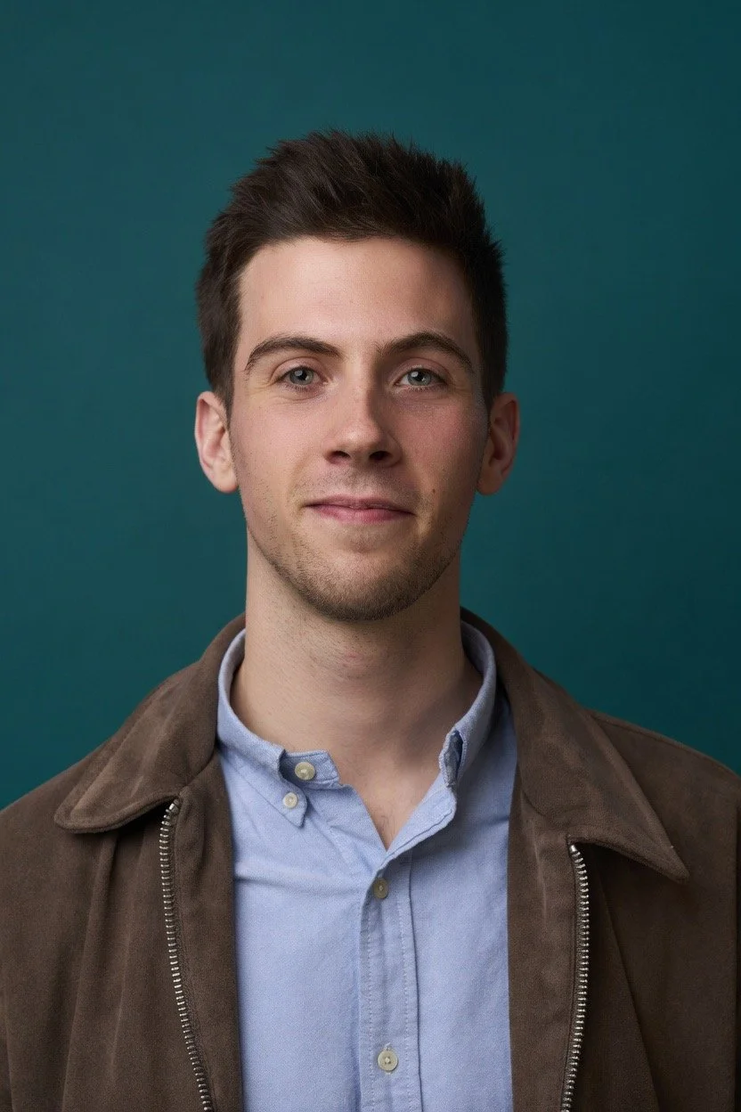 Portrait of Lewis with short dark hair, wearing a light blue button-up shirt and a brown jacket, standing against a teal background.