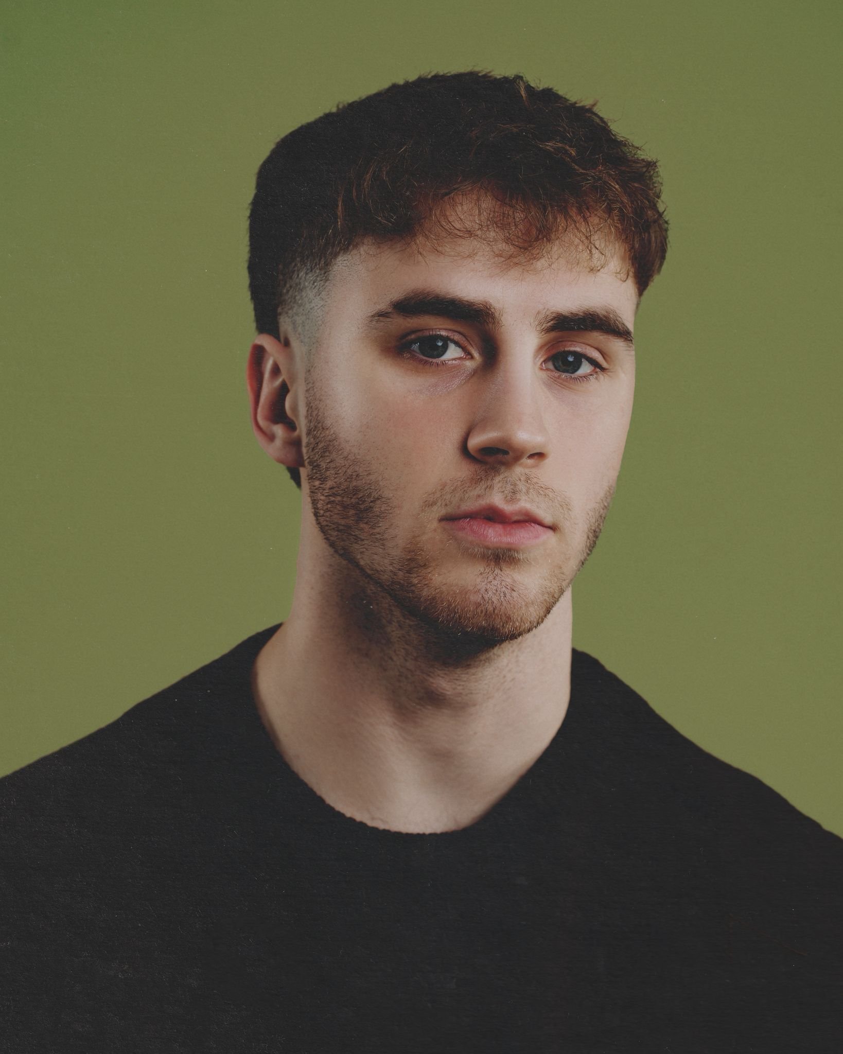 Headshot of Angus with dark, slightly curly hair, blue eyes, light skin, and a black shirt, against a green background.