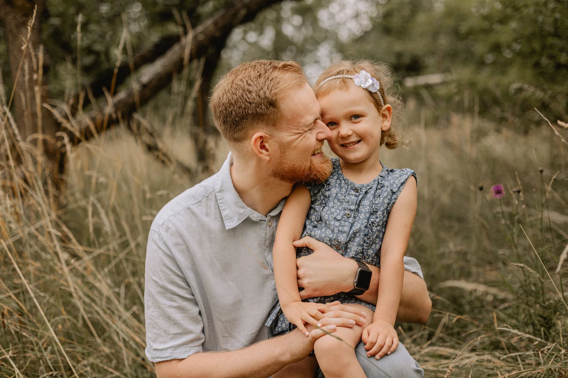 Ein Mann mit rotem Bart und weißem Hemd hält ein lachendes Mädchen mit Blumenhaarband in einem Feld mit hohem Gras und Bäumen im Hintergrund.