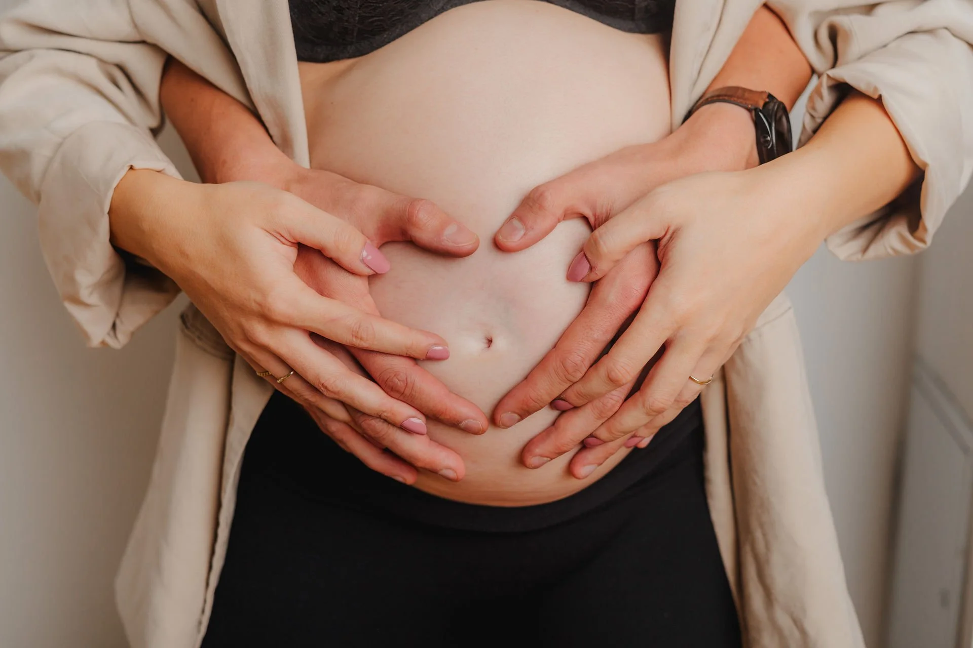 Eine schwangere Person liegt mit offenen Oberkörper, während mehrere Hände ein Herz auf ihrem Bauch formen.