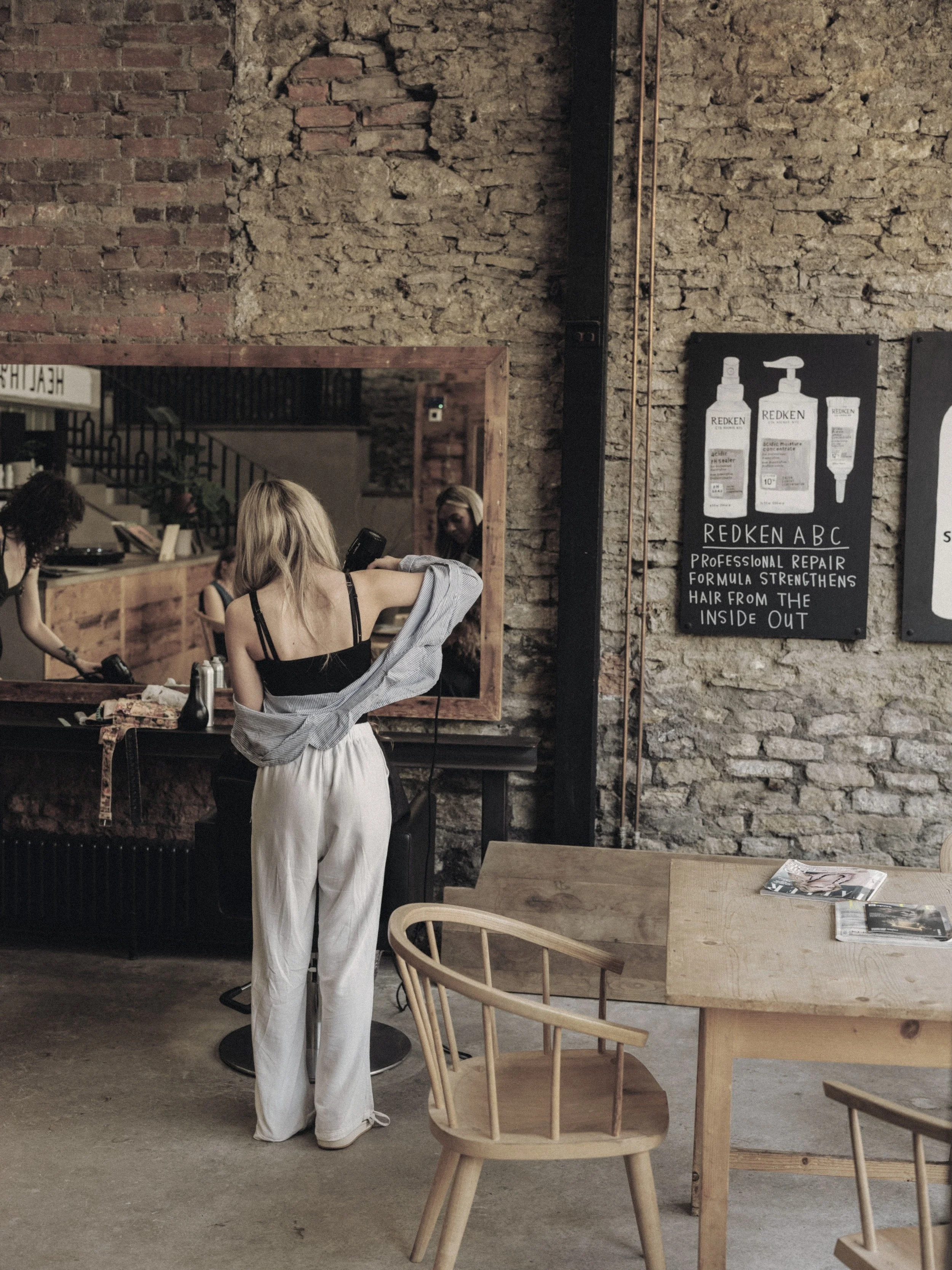 A woman with long blonde hair standing in a salon, getting her hair cut or styled by a barber in front of a large mirror. The salon has an exposed brick wall with a red and black sign advertising Redken hair products.