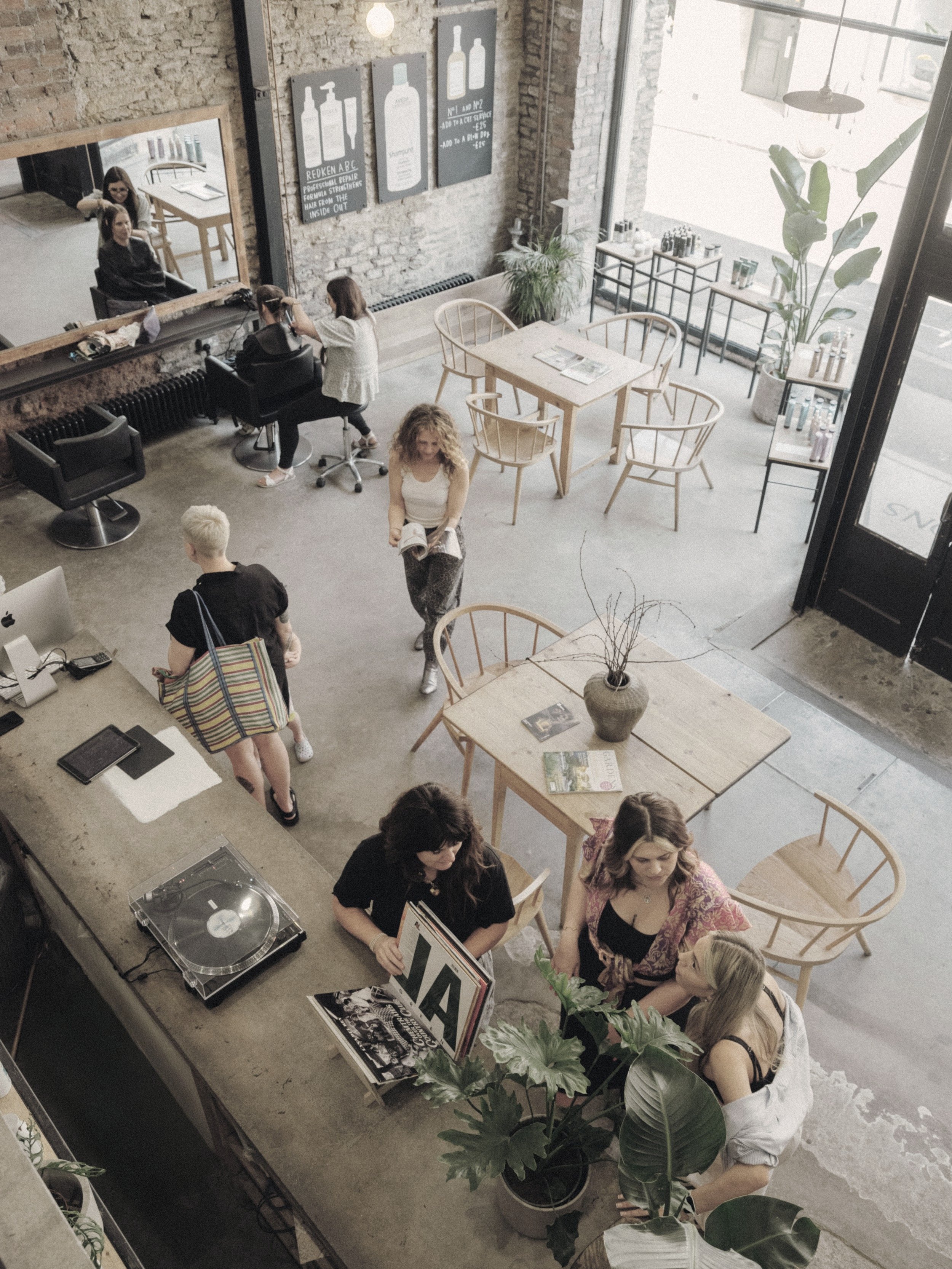 People inside a modern, stylish cafe with brick walls, large windows, and minimalistic wooden furniture. Some are sitting at tables; others are standing or walking. The space features a record player, vinyl records, plants, and wall art.