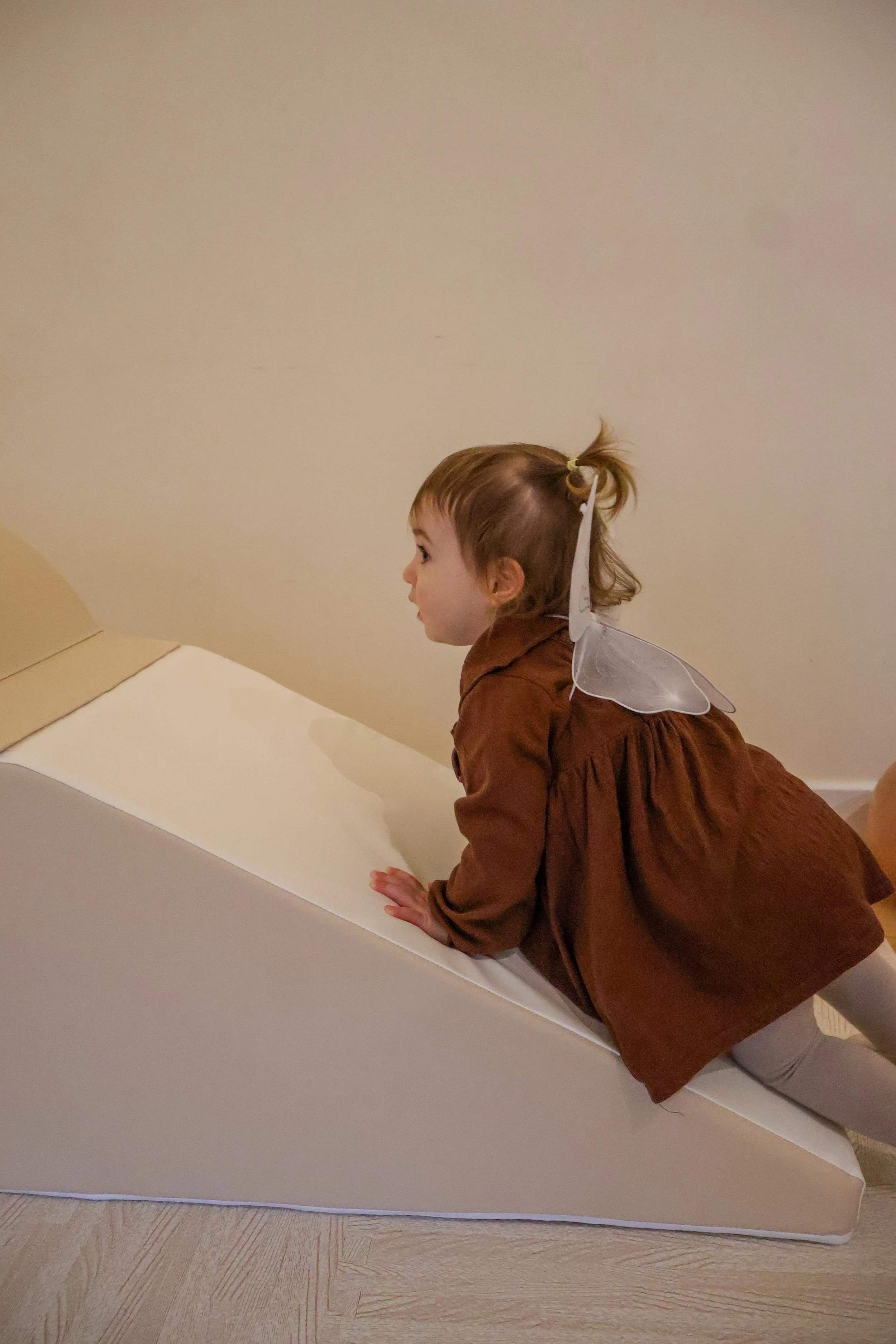 Toddler wearing angel wings climbing a soft ramp indoors.