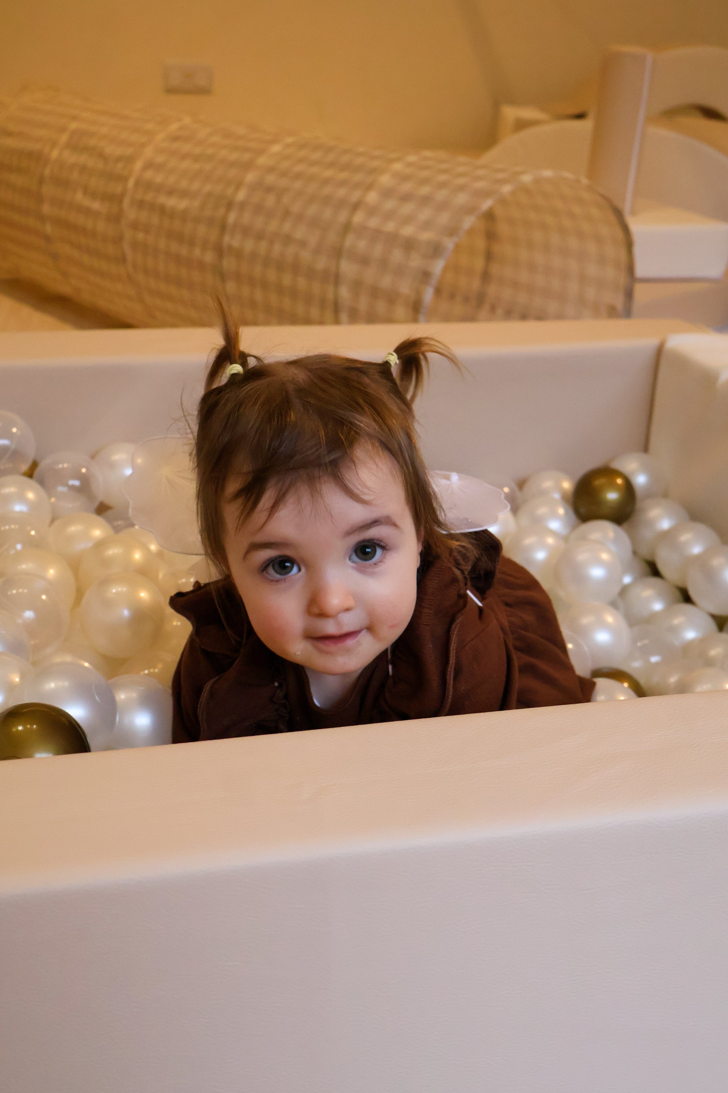 A toddler with pigtails playing in a ball pit filled with white and gold balls.