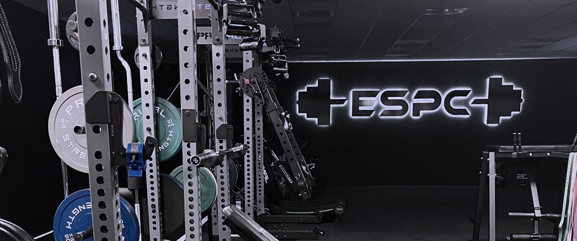 Gym equipment including weight plates, a squat rack, and a structured sign with the word 'ESPEC' on a black wall.