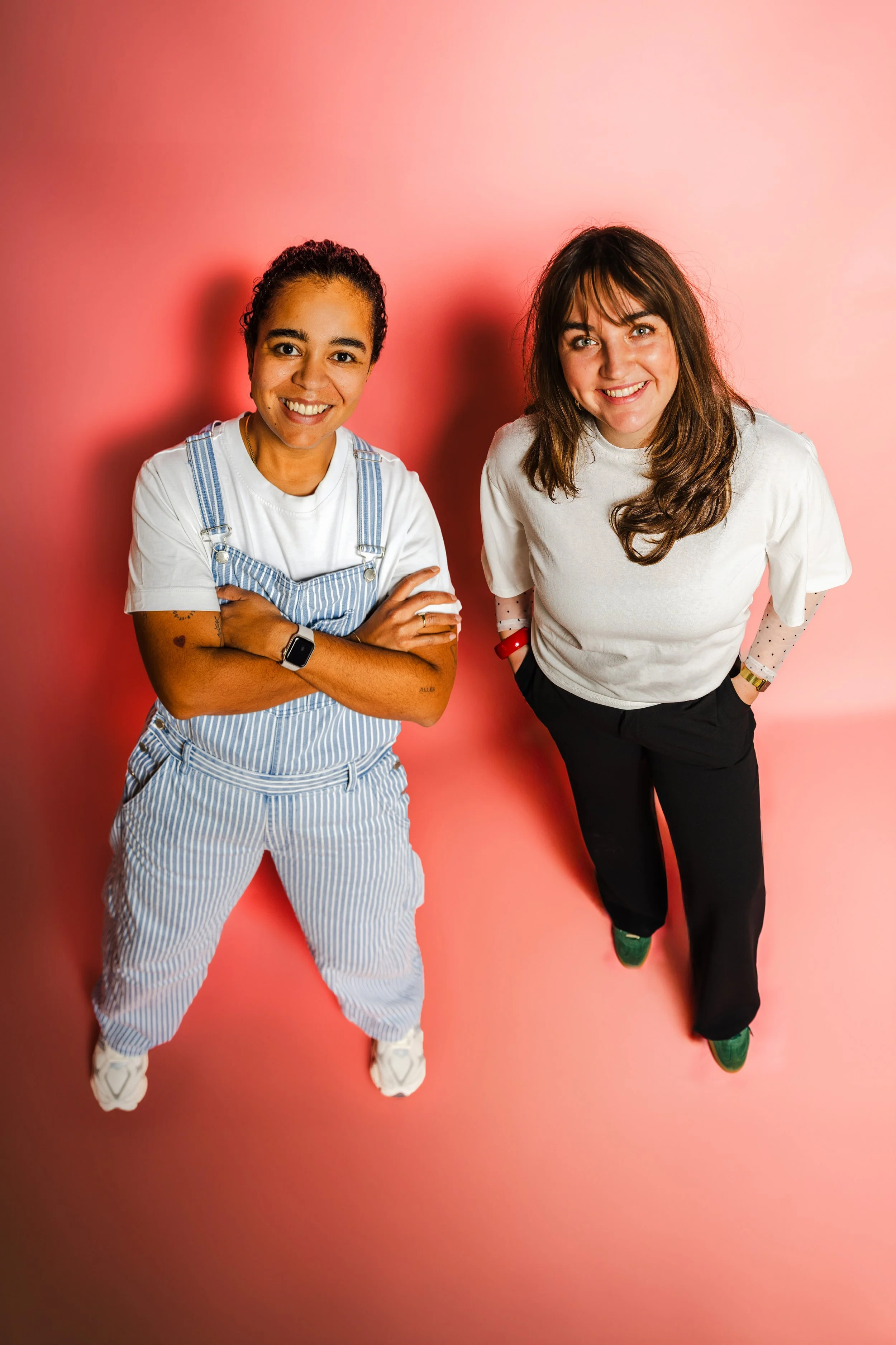 Twee vrouwen lachen en poseren tegen een roze achtergrond.