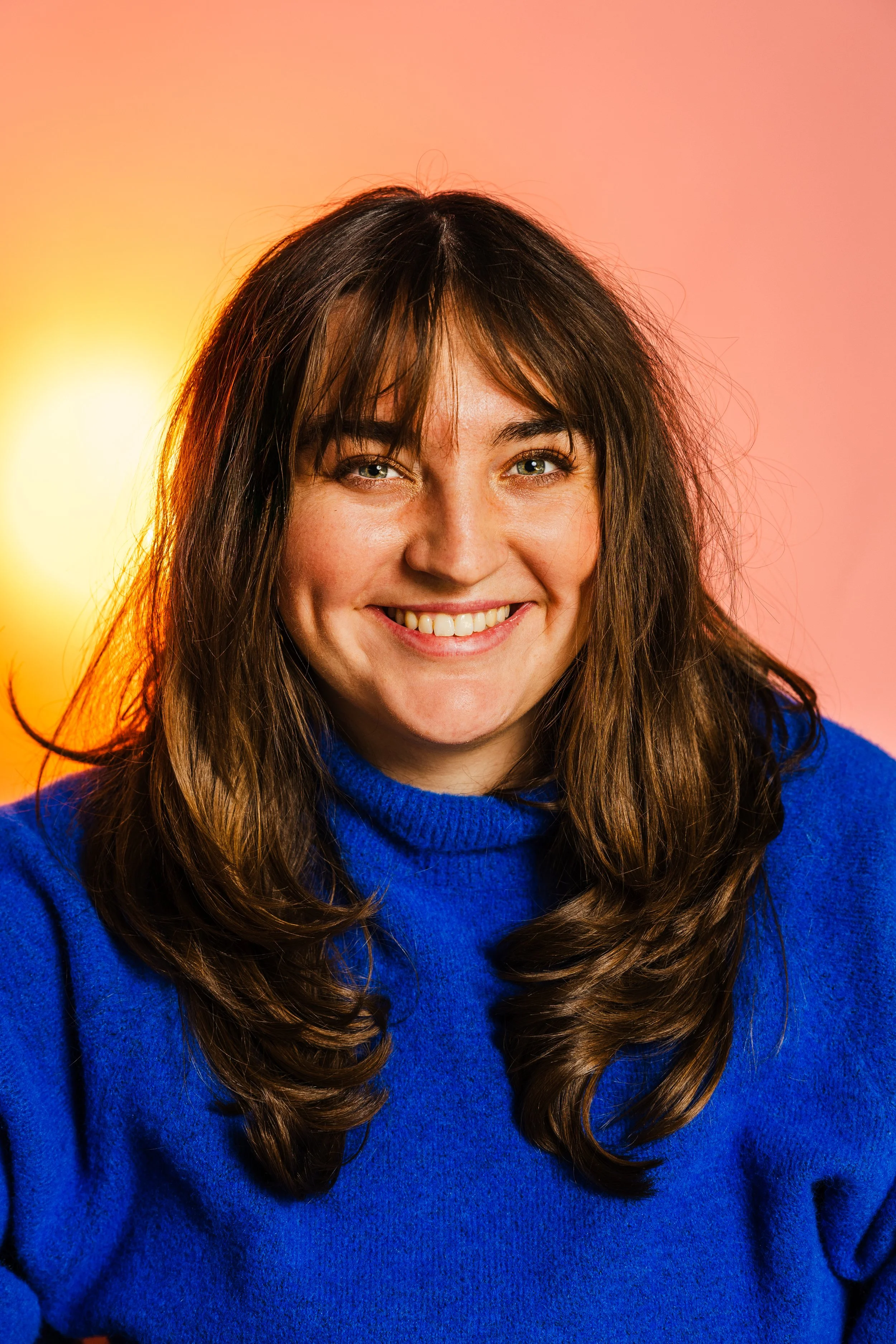 A woman with long, wavy brown hair smiling at the camera against a light pink background.