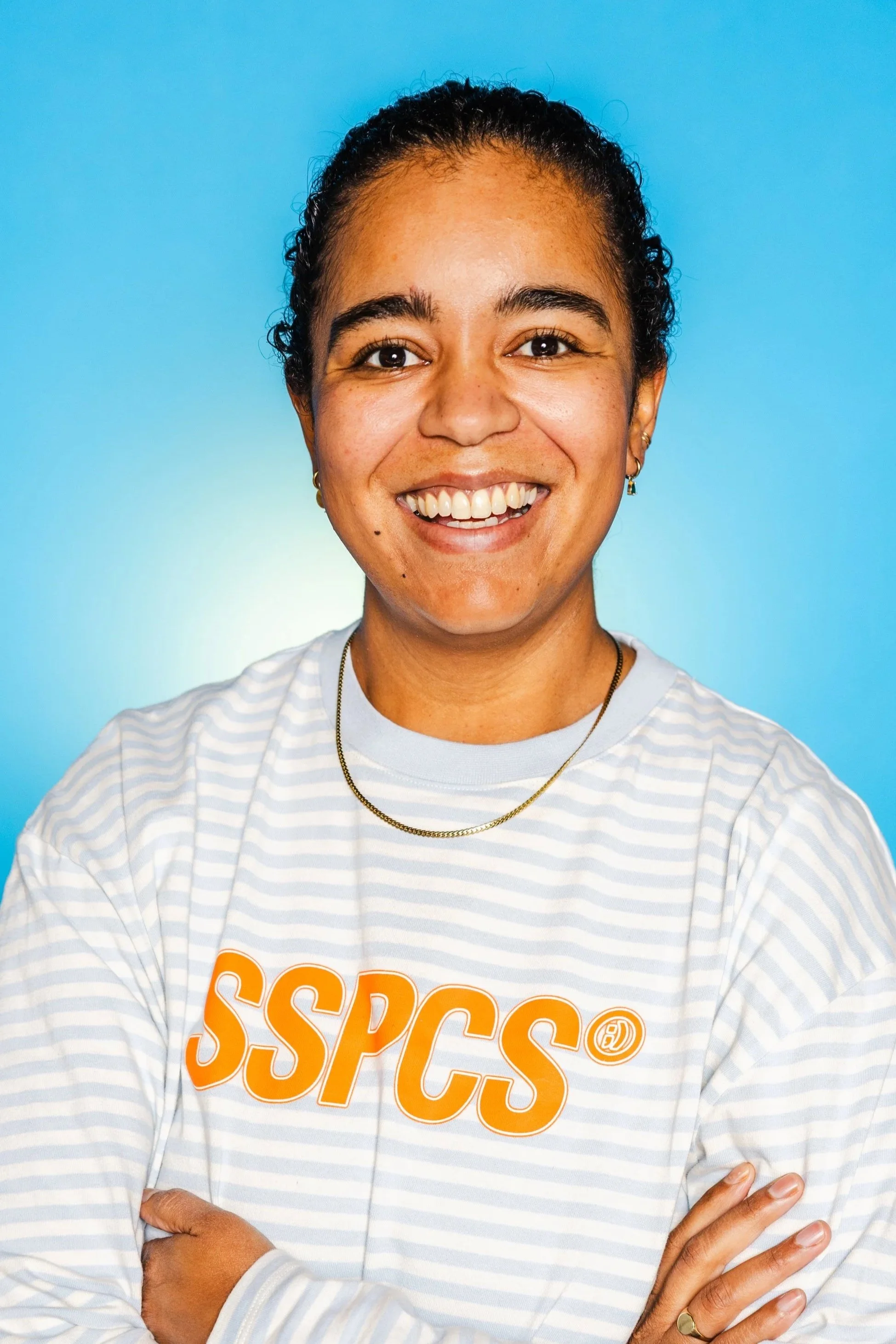 A smiling woman with dark hair in a bun, wearing a white shirt and black overalls, standing against a light blue background.