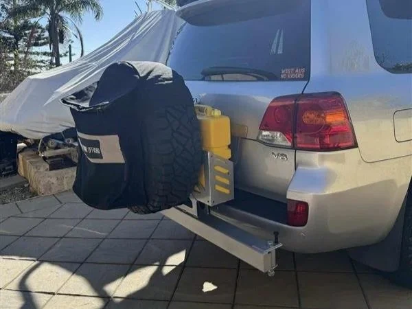 Silver car with a mounted spare tire, yellow gas tank, and a sliding metal rail system at the rear, parked on tiled pavement with a covered vehicle and trees in the background.