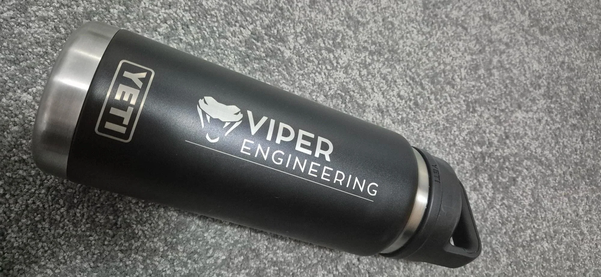 A black stainless steel water bottle with a silver top, lying on a gray carpet. The bottle features a logo with a stylized snake and the words "VIPER ENGINEERING" on it.