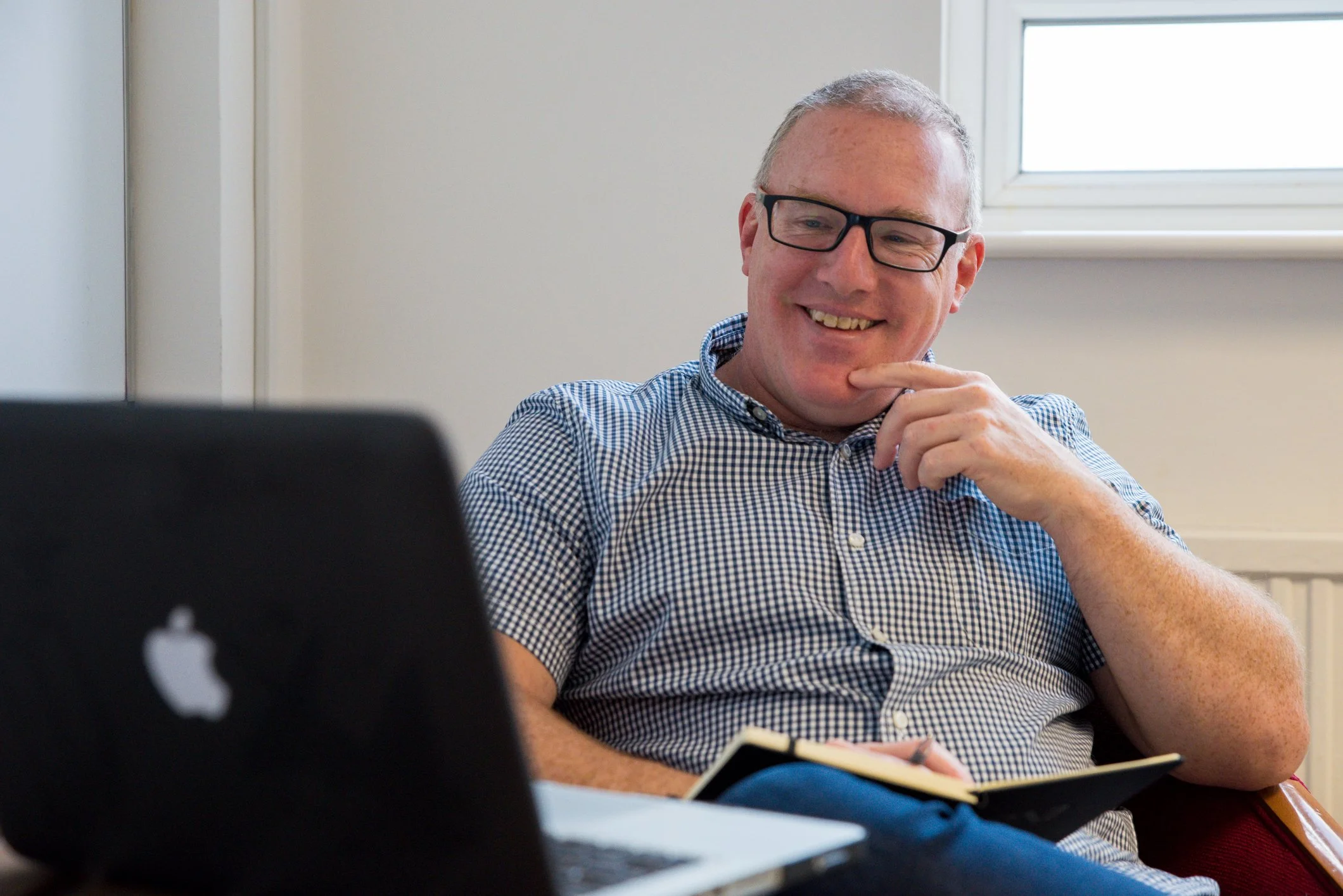 A man with glasses and a checkered shirt sitting at a table, smiling and looking at a laptop.