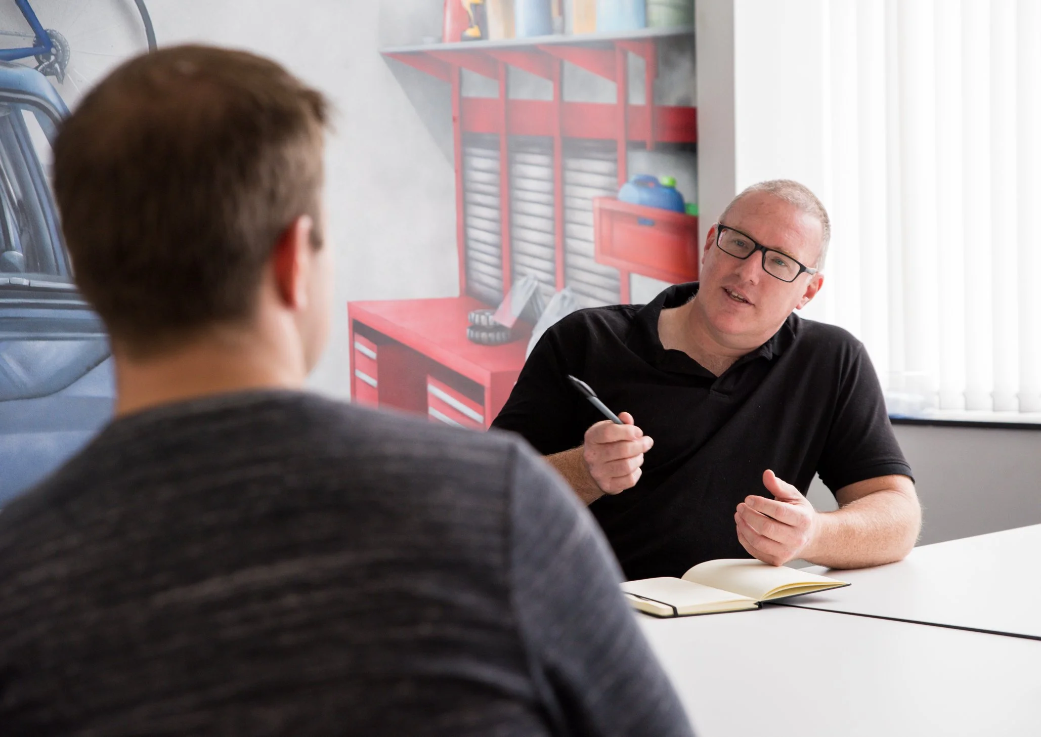 Two men are sitting at a table in a discussion. One man, with glasses and short hair, is speaking and holding a pen. The other man, seen from behind, has short brown hair. There is a yellow notebook open on the table.
