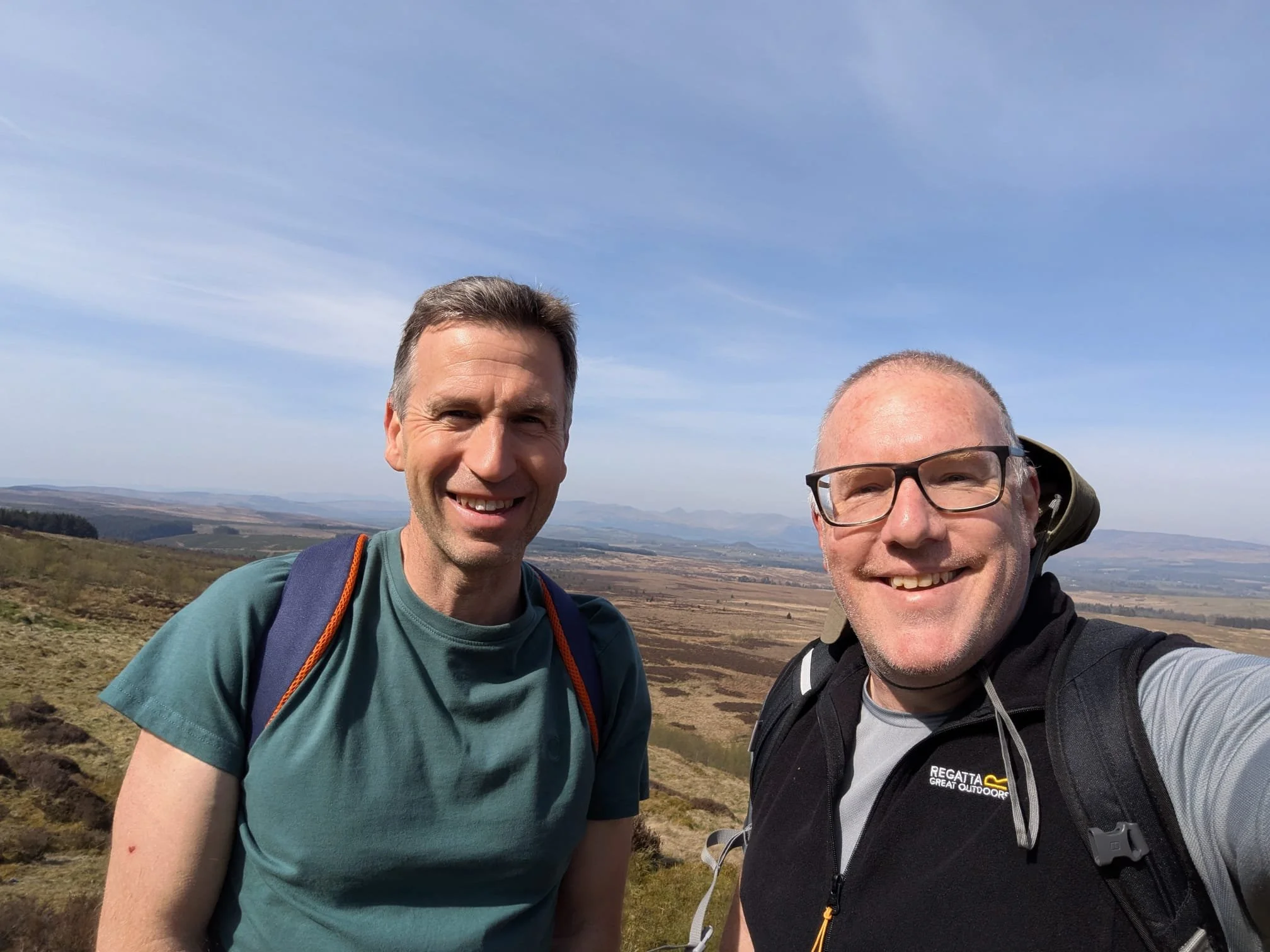 Two men smiling outdoors in a hilly landscape with a clear blue sky, taking a selfie together with the horizon stretching behind them.