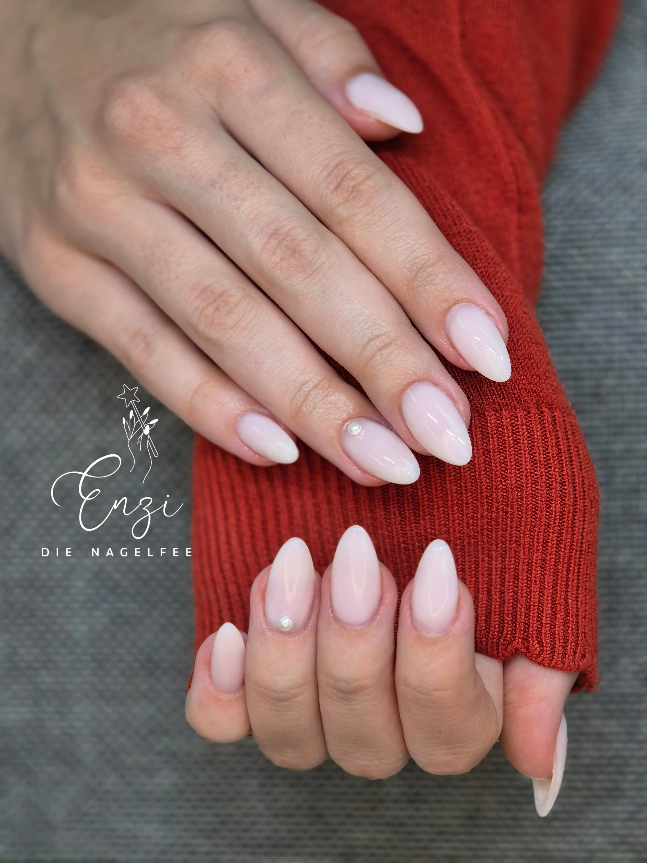 Nahaufnahme von gepflegten Fingernägeln mit lackiertem, glänzendem, nudefarbenem Nagellack und kleinen Perlendekorationen, gehalten vor einem grauen Stoff und rot gestricktem Kleid.