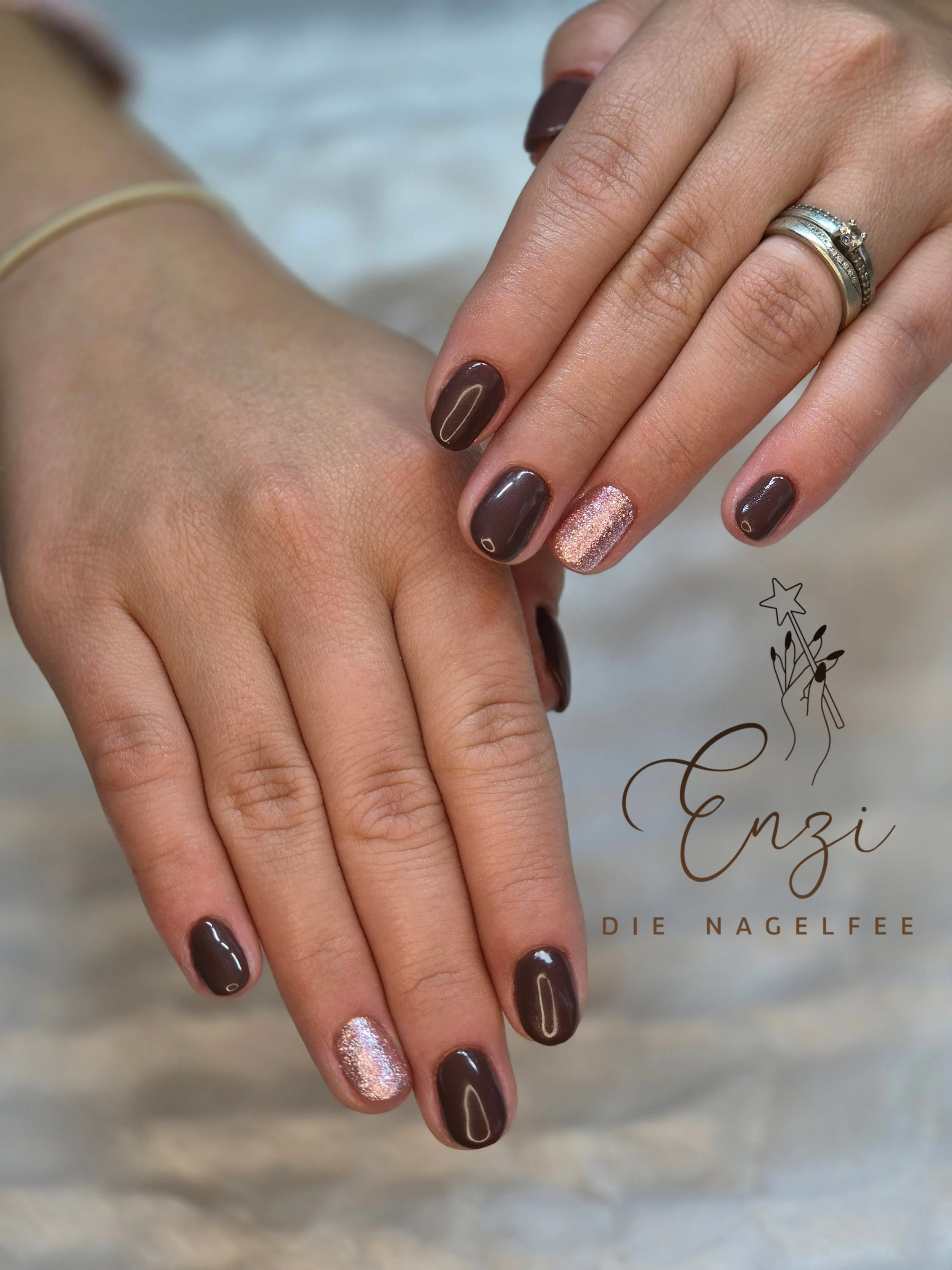 Nahaufnahme von Händen mit braunen und glitzernden pinken Nagellacken und Ringen, auf einem weißen Stoffhintergrund.