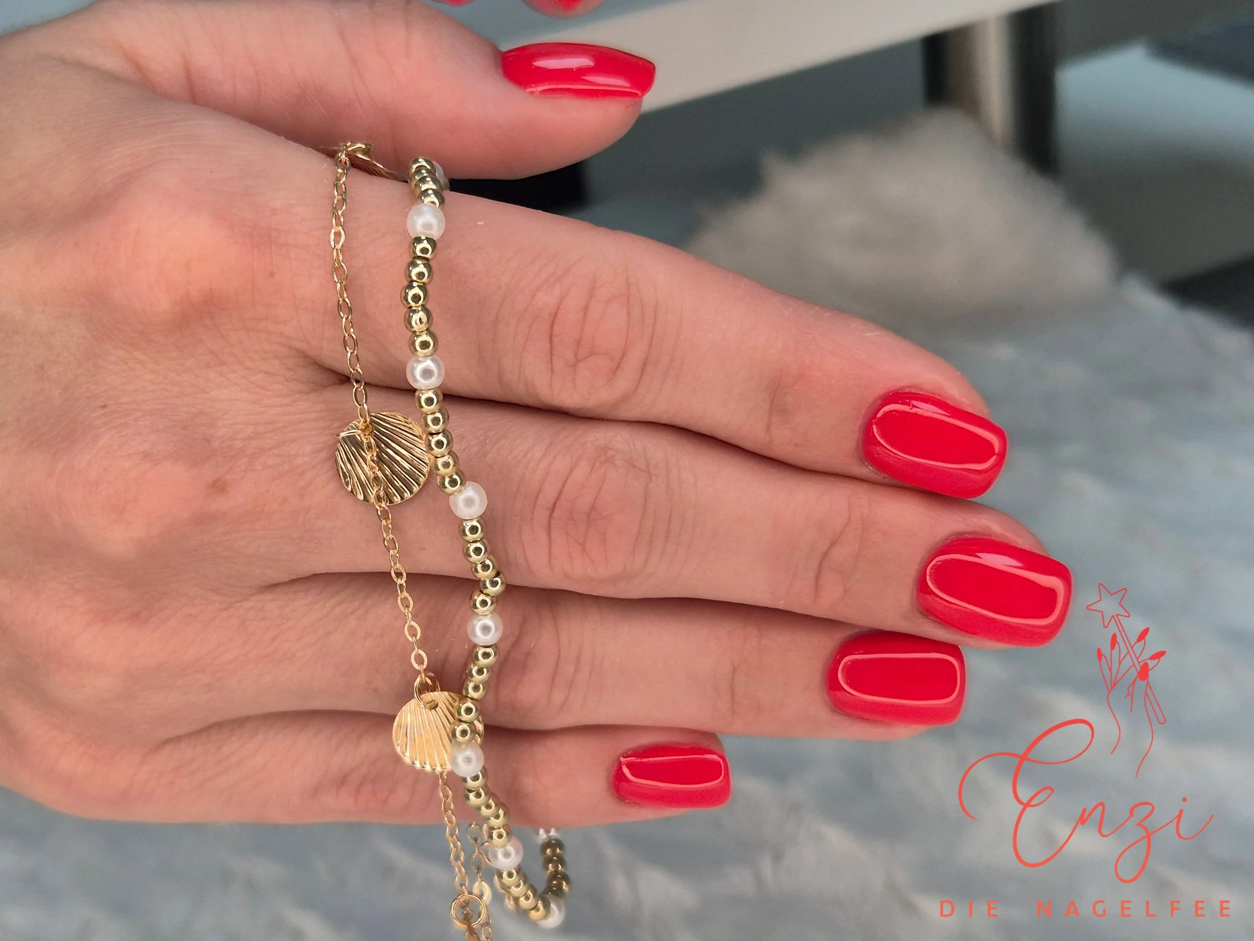 Nahaufnahme einer Hand mit roten Nägeln, die mehrere goldene Schmuckketten mit Muschelanhängern und Perlen hält.