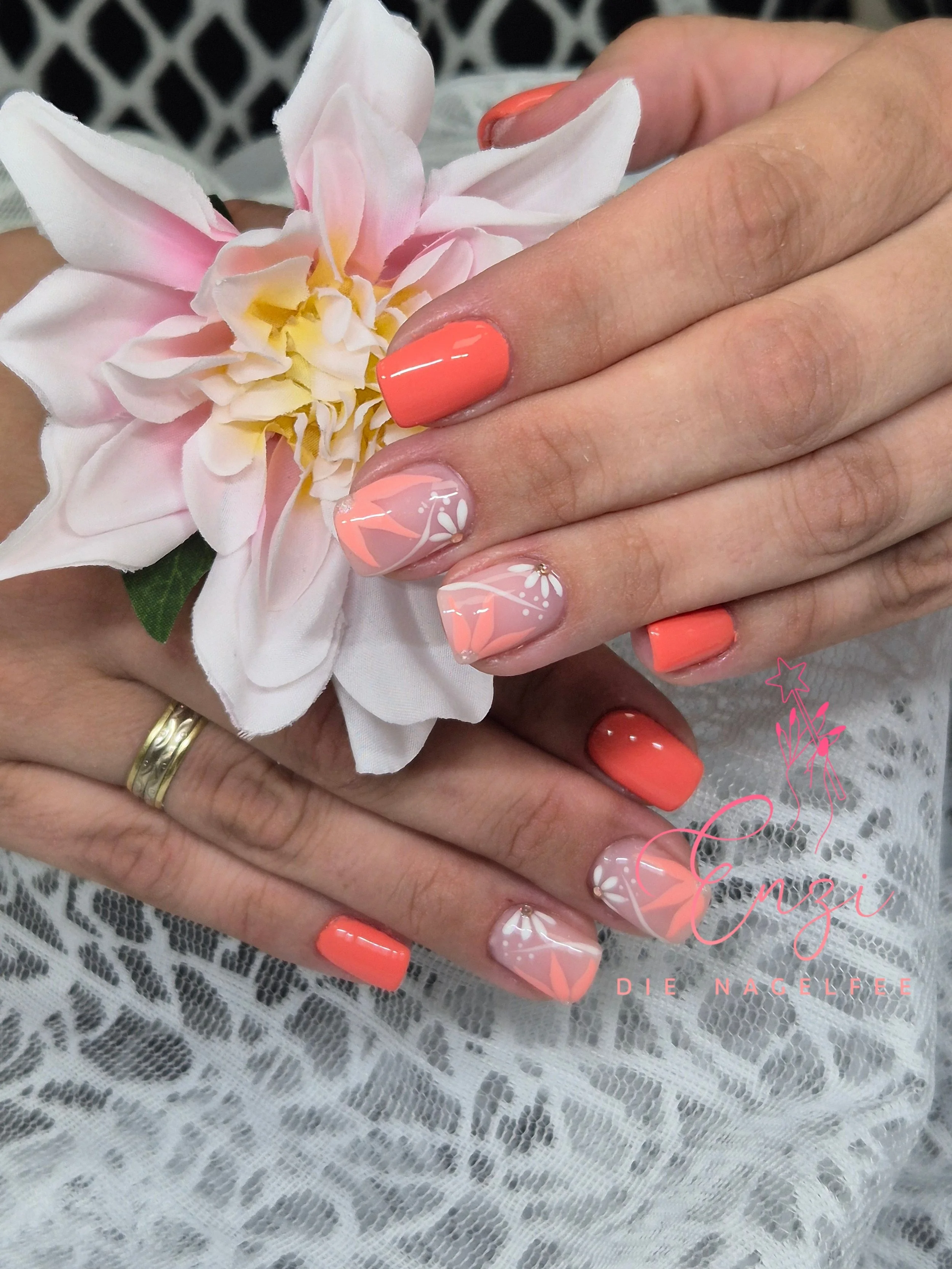 Hände mit leicht gebräunten Nägeln in Korallenfarbe, die auf einer Spitze mit weißen und pastellrosa Nageldesigns liegen, halten eine große, pink-weiße Blume.